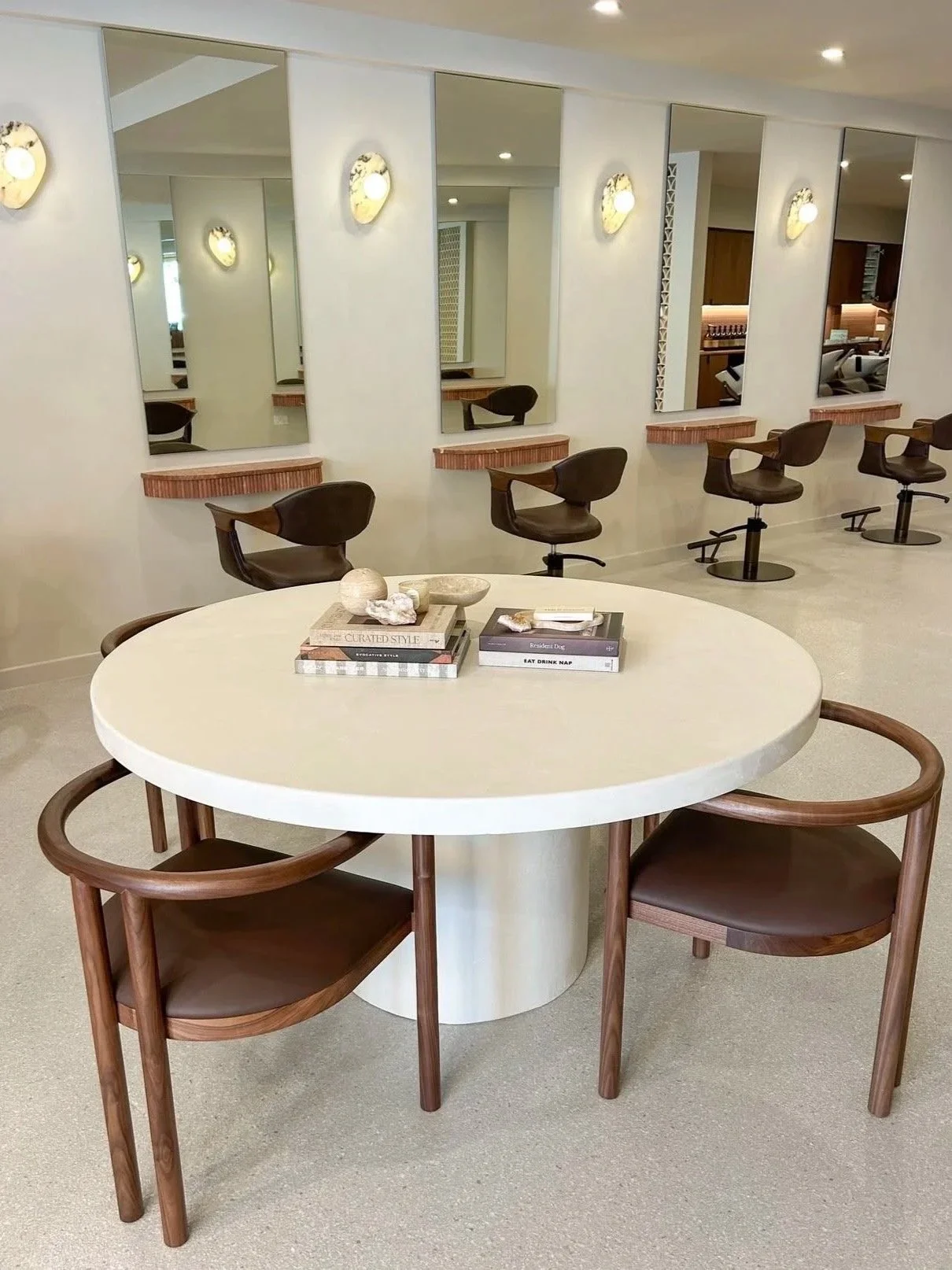 Modern hair salon with a round table in the foreground, decorated with books and decorative items. In the background, there are five styling chairs in front of mirrors on a white wall, illuminated by wall-mounted lights.