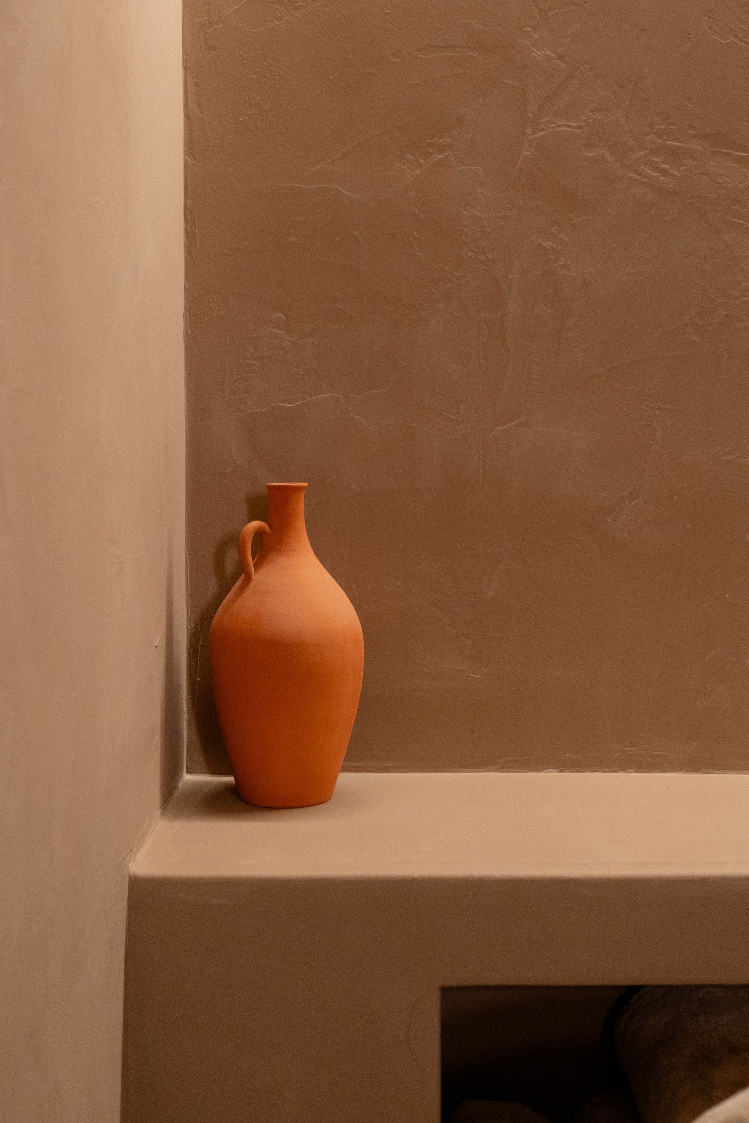A reddish-brown ceramic vase with a small handle on a white shelf, against a background of a textured brown wall and a beige wall.