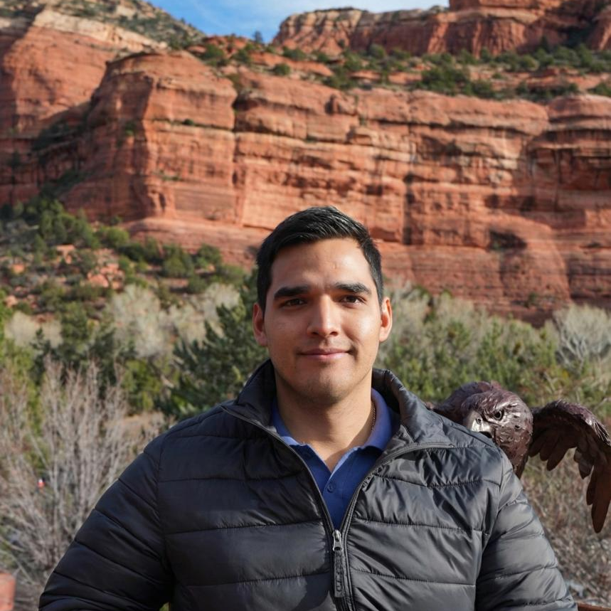 A young man standing outdoors with red rock cliffs in the background and a bird perched on his shoulder.