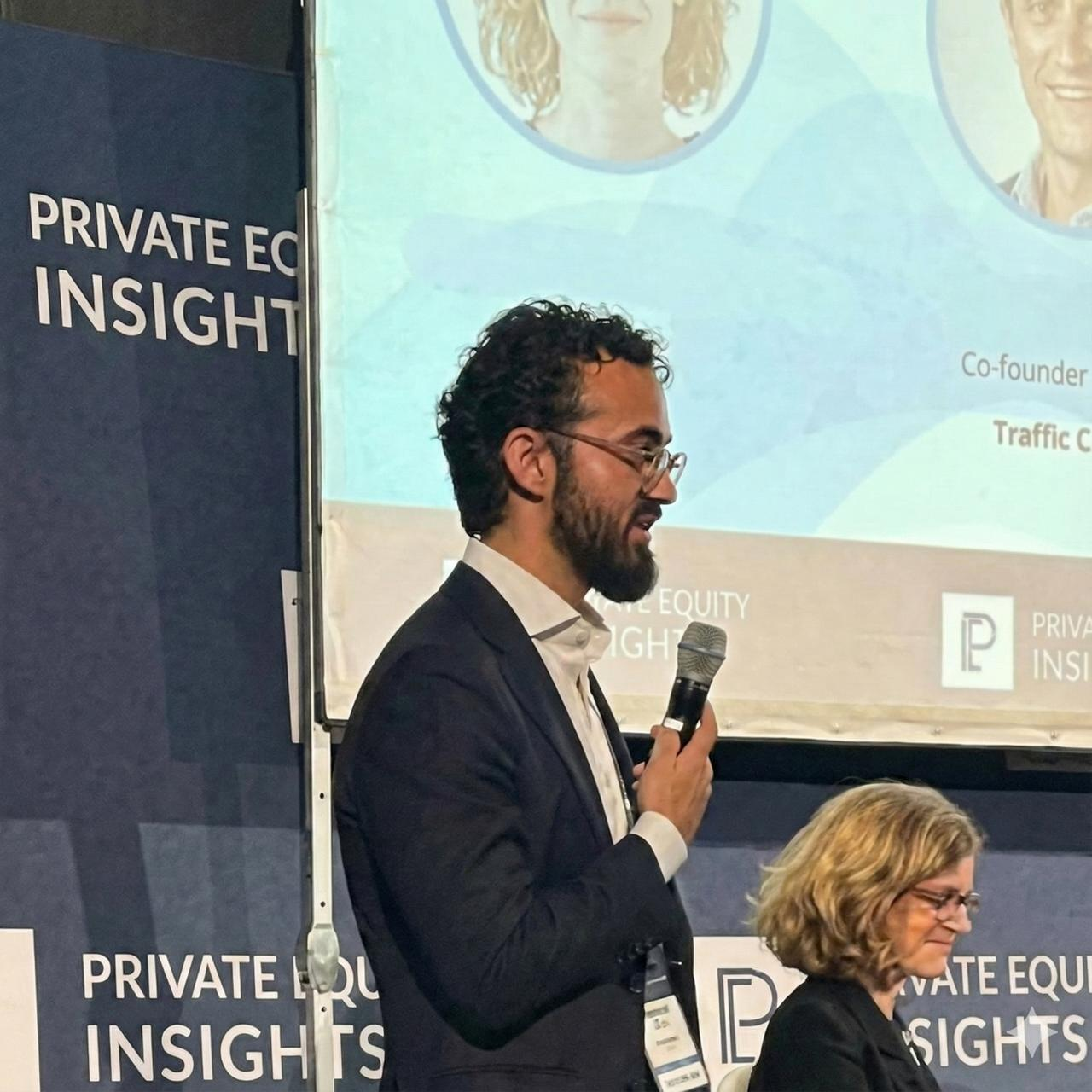 A man with curly dark hair and a beard, dressed in a dark suit and white shirt, holding a microphone and speaking. A woman with glasses and shoulder-length blond hair, dressed in black, is seated beside him, looking down. They are at a conference with a backdrop that reads 'Private Equity Insights' and a screen presenting their names and titles.