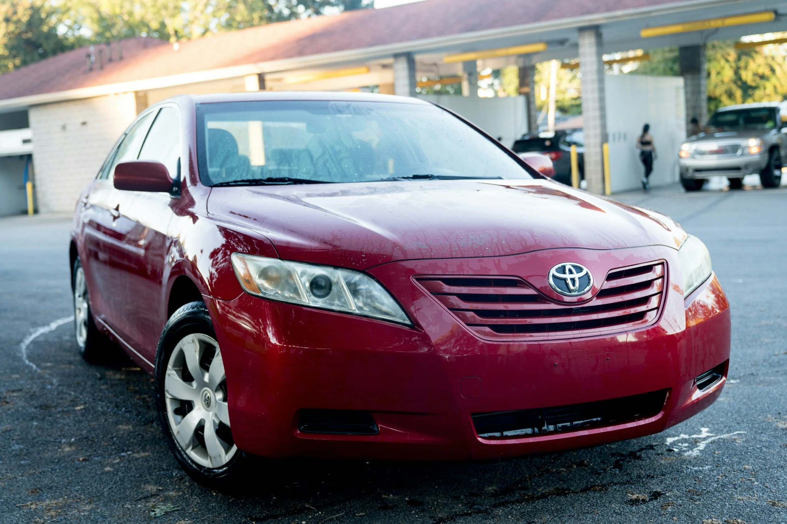 Red Toyota Camry parked in a parking lot with a building and other vehicles in the background.