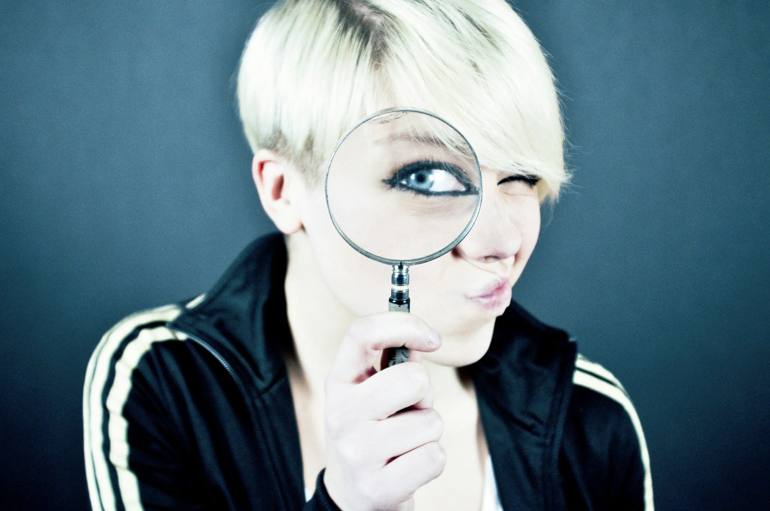 Person with short blonde hair holding a magnifying glass over one eye, squinting and pursing lips, wearing a black jacket with white stripes against a dark background.