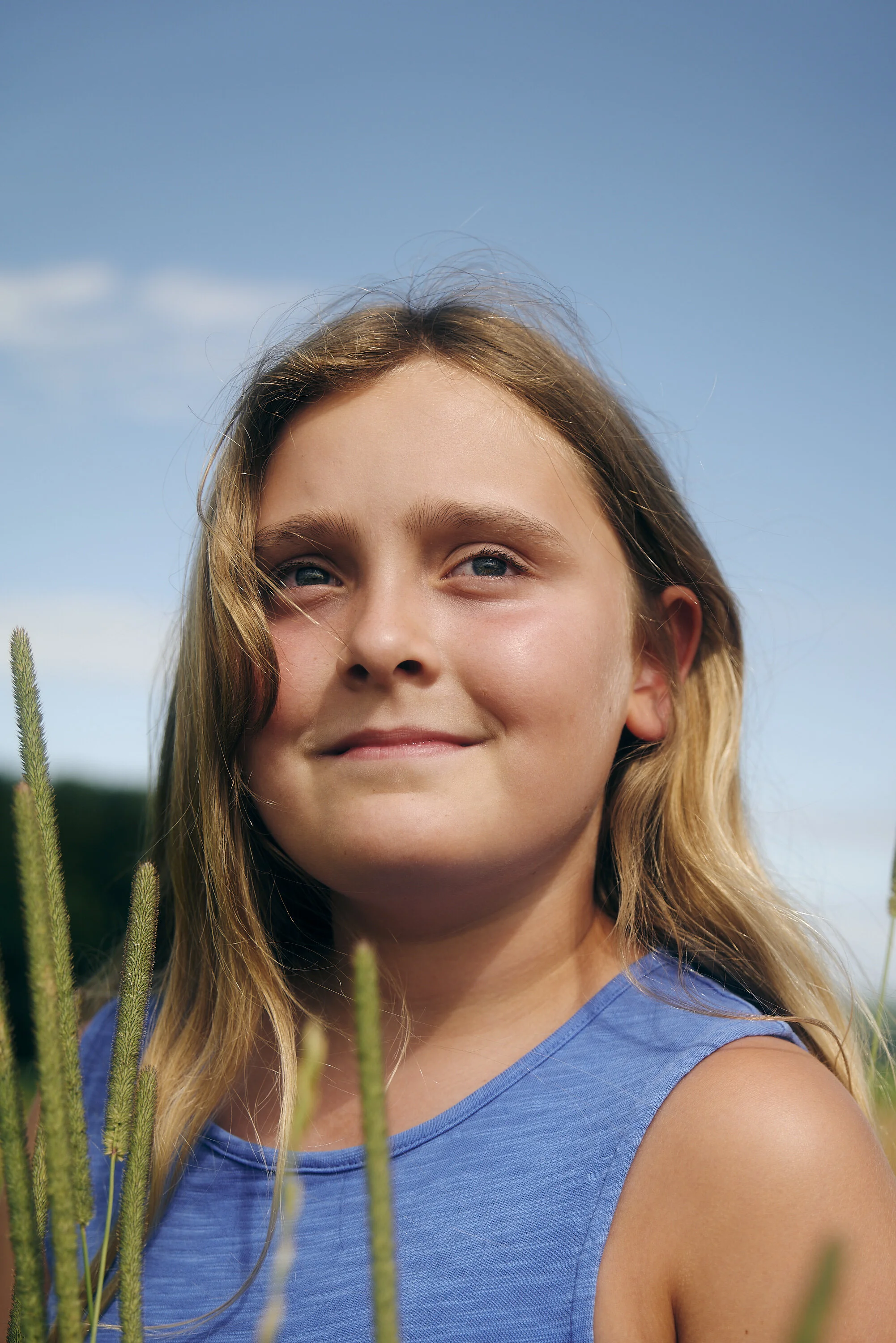 Porträt eines Mädchens mit blondem Haar im Freien, das in die Ferne schaut. Im Vordergrund sind Grashalme zu sehen. Der Himmel ist klar und blau.