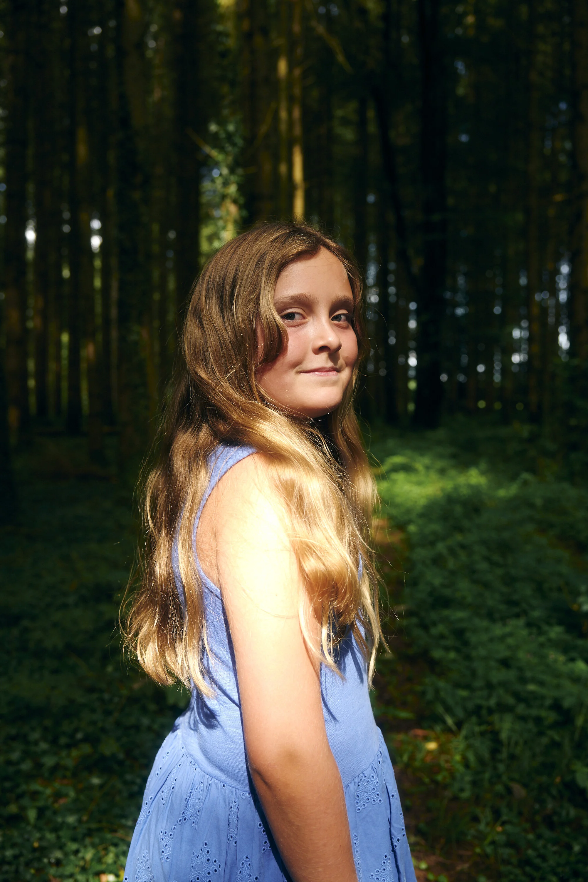 Mädchen in einem blauen Kleid steht in einem Wald, Sonnenlicht fällt auf sie.