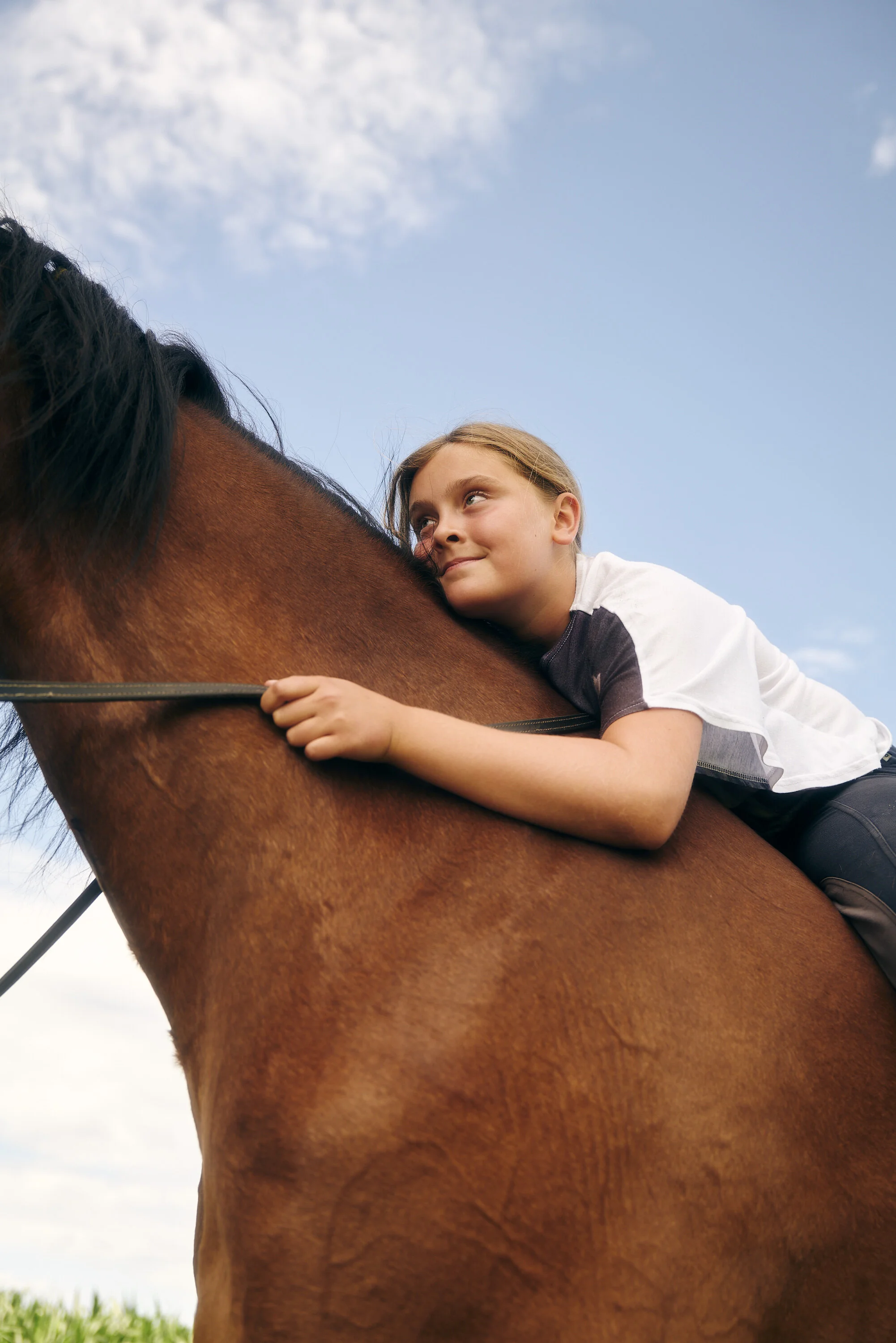 Ein Kind reitet auf einem braunen Pferd und schaut nach oben in den Himmel. Der Himmel ist blau mit einigen Wolken.