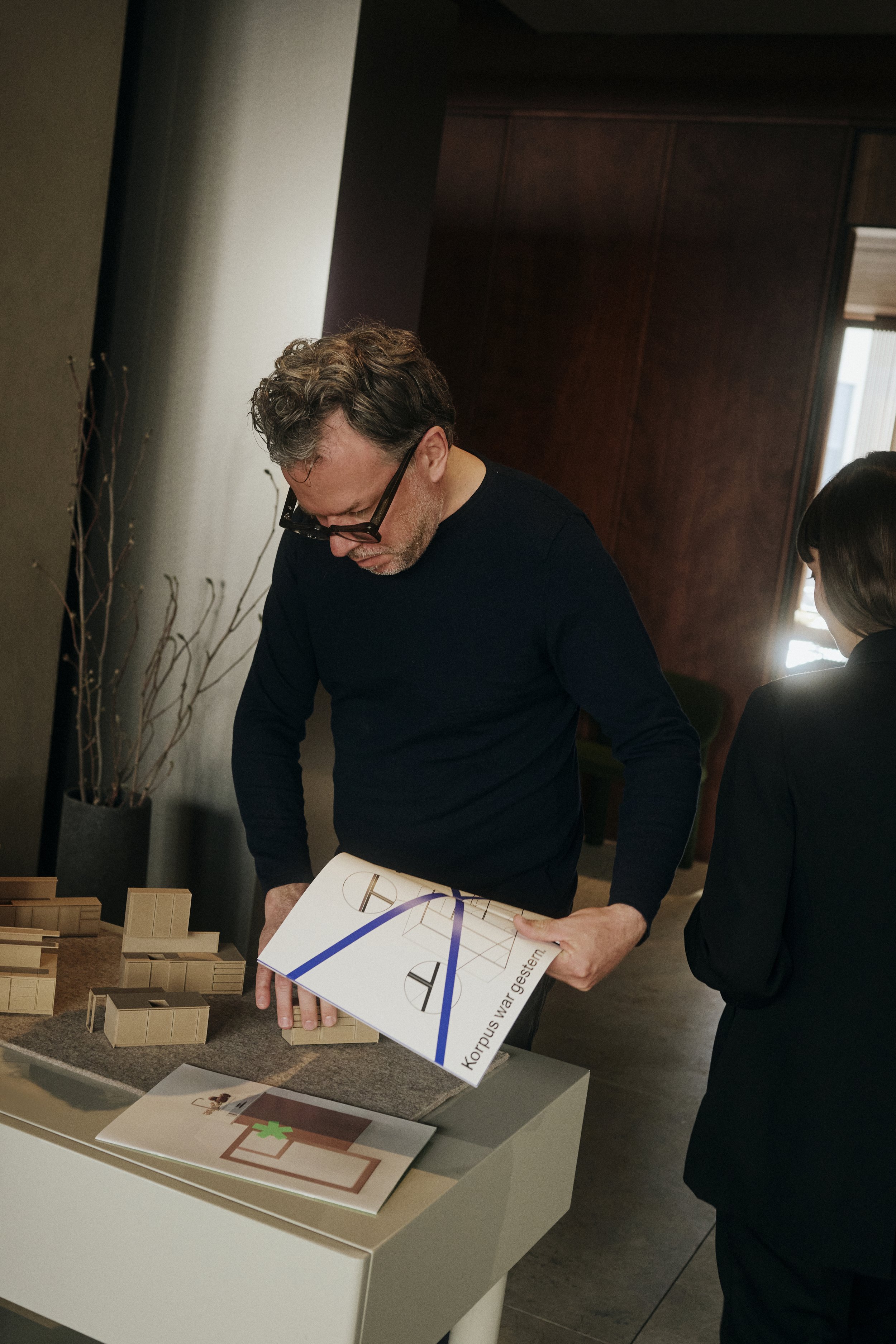 Mann mit Brille betrachtet ein Architekturmagazin, Modell eines Gebäudes auf einem Tisch, Person im Hintergrund.