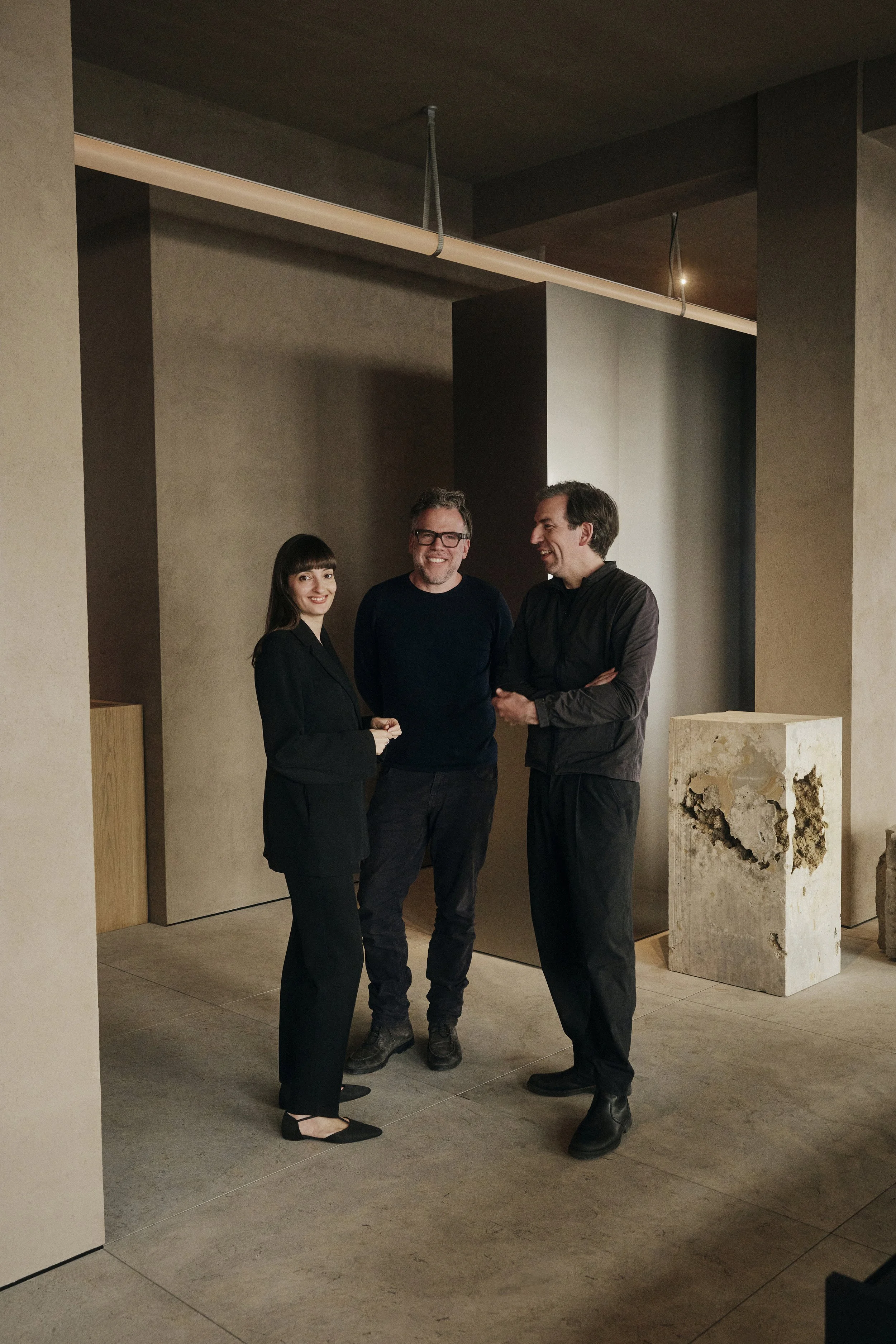 Drei Personen stehen in einem minimalistischen, modernen Innenraum. Sie tragen dunkle, legere Kleidung und lächeln oder unterhalten sich. Im Hintergrund sind künstlerische Wandelemente und moderne architektonische Details zu sehen.