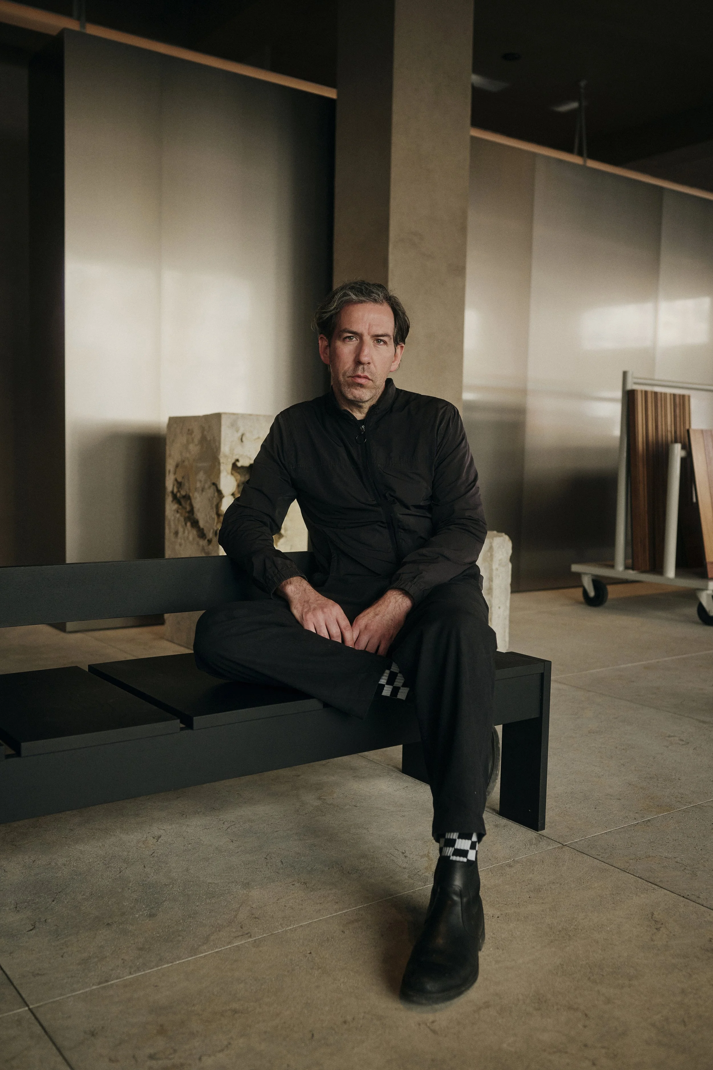 Eine Person sitzt auf einer schwarzen Bank in einem modernen Innenraum, umgeben von minimalistischen Designmöbeln und Architekturmaterialien, wie Steinquadern und Holzplatten auf Rollwagen. Die Person trägt ein schwarzes Outfit mit karierten Socken u