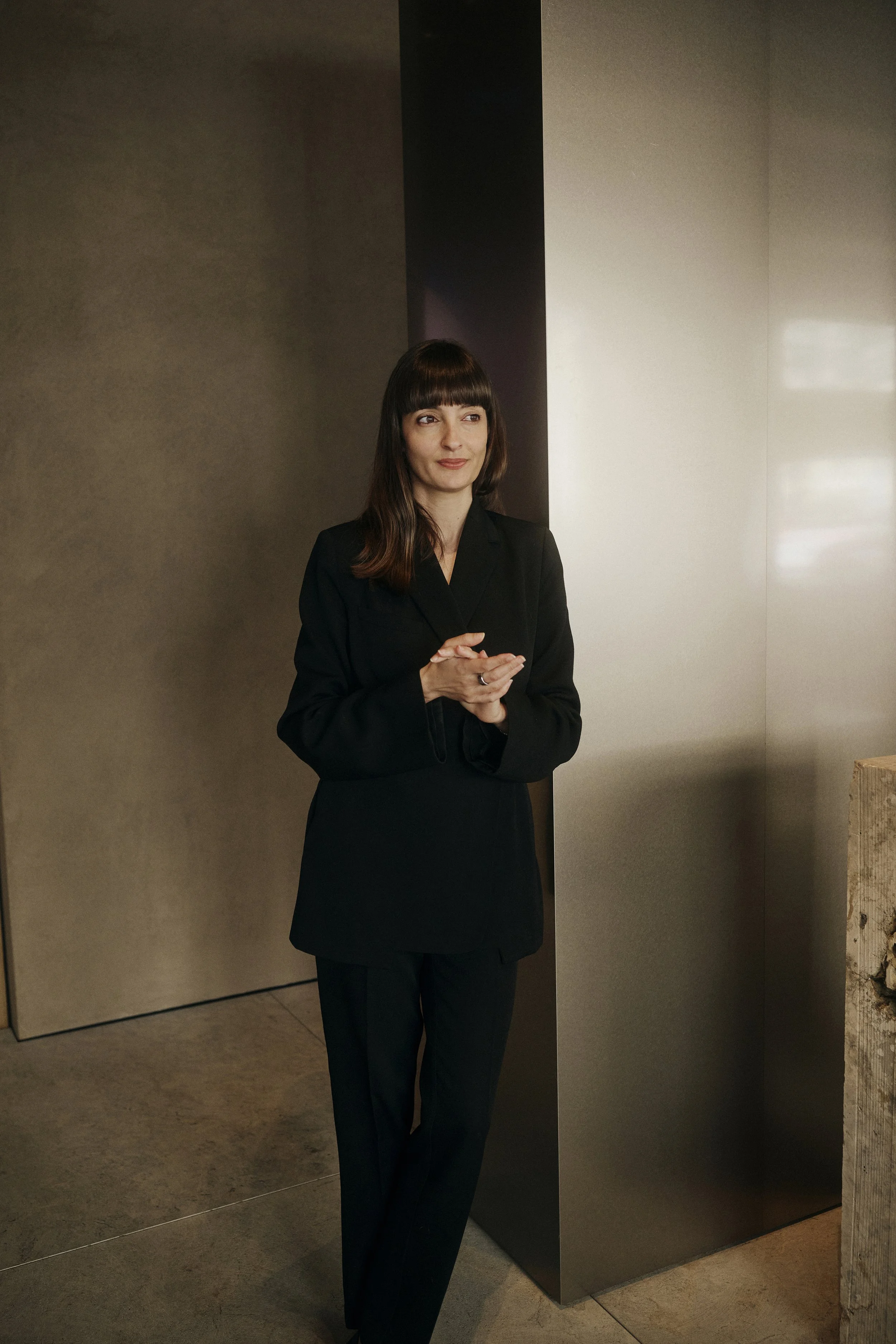 Frau in schwarzem Anzug steht in modernem Innenraum, Hände gefaltet, neutraler Hintergrund.