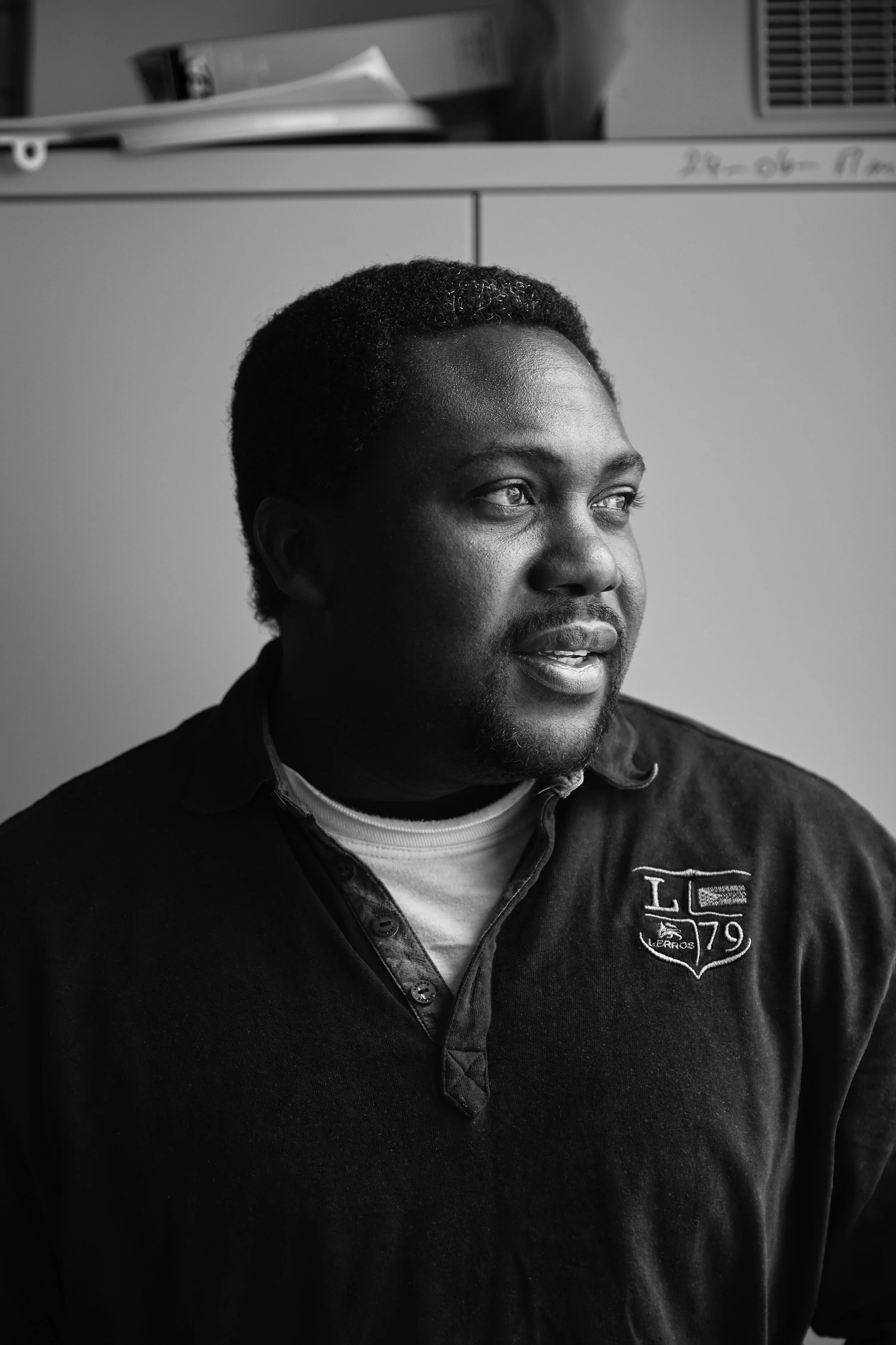 Schwarz-Weiß-Foto eines Mannes, der nach rechts schaut, mit kurzem, lockigem Haar und einem Poloshirt, im Hintergrund ein Aktenschrank und ein Regal mit Dokumenten.