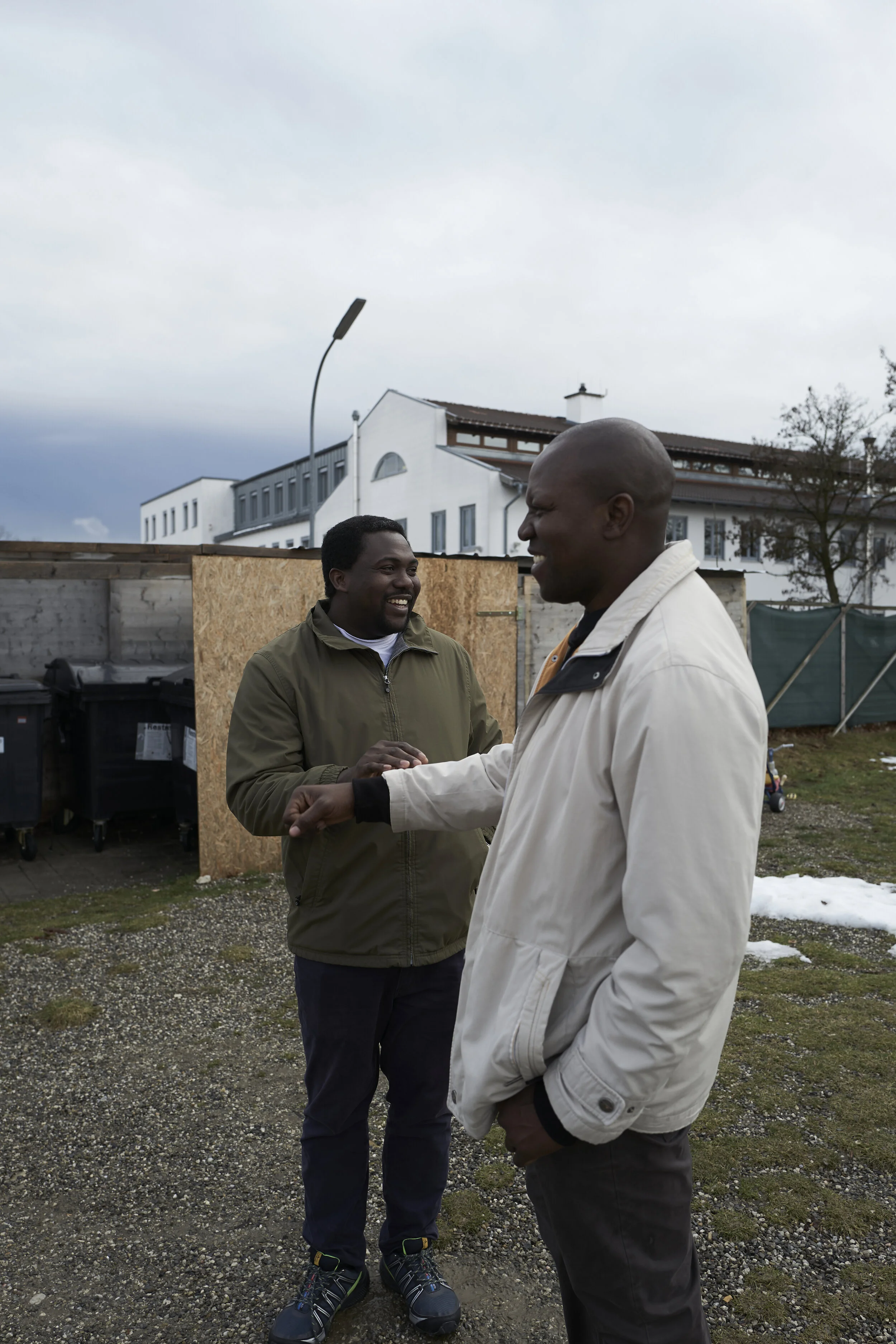 Zwei Männer unterhalten sich draußen bei bewölktem Himmel; einer trägt eine grüne Jacke, der andere eine weiße Jacke. Im Hintergrund sind Gebäude und Mülltonnen zu sehen.
