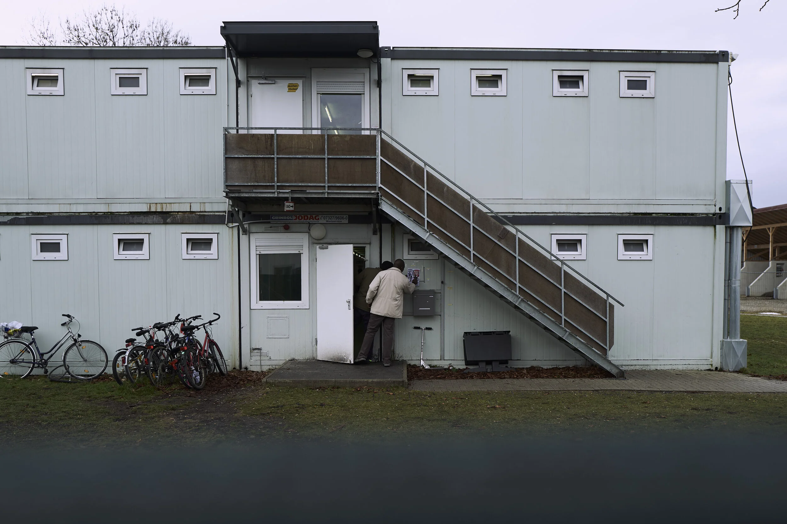 Zwei Personen stehen vor einem grauen, zweistöckigen Containergebäude mit einer Außentreppe. Mehrere Fahrräder sind links neben dem Eingang geparkt.