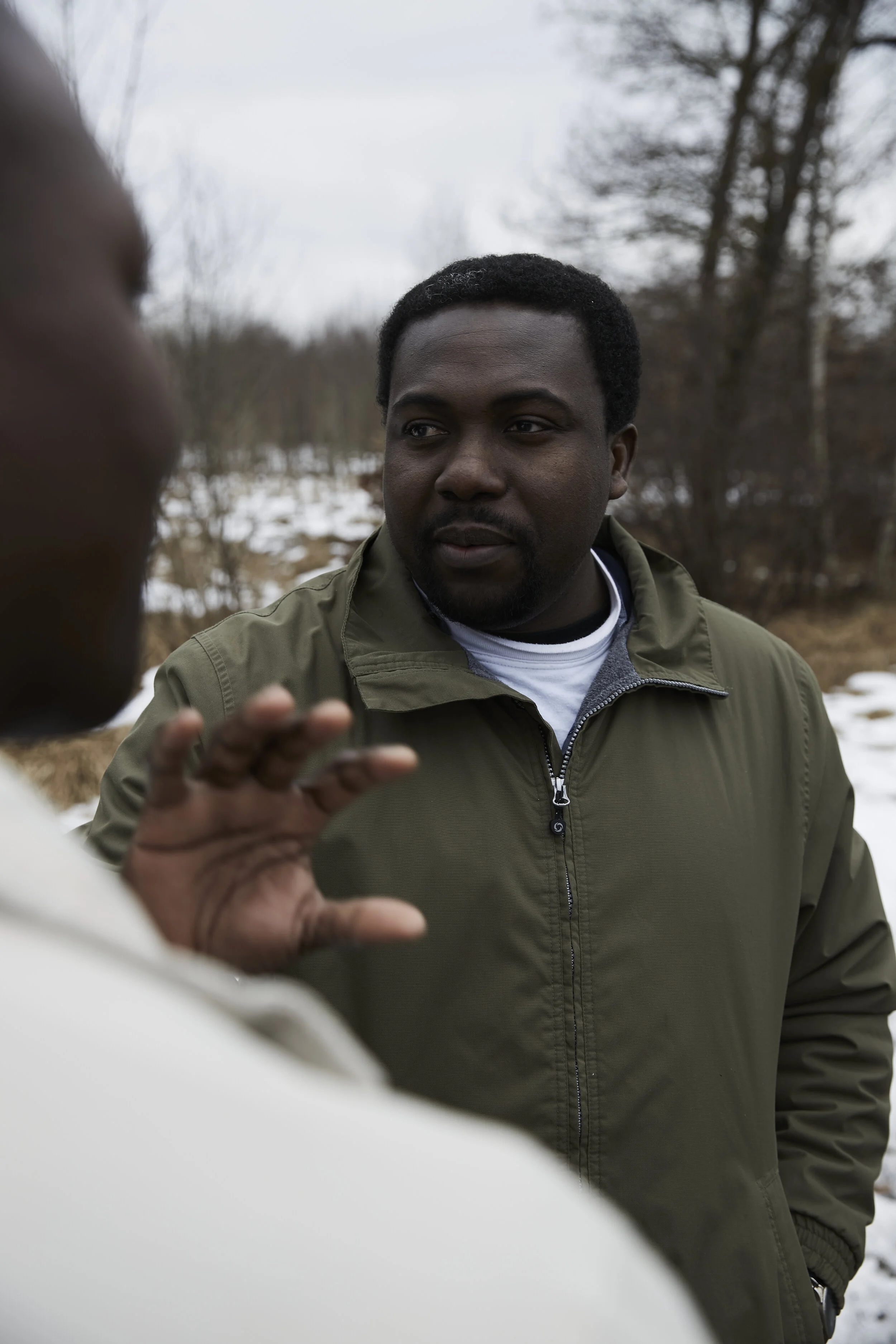 Zwei Personen im Freien im Gespräch, eine Person in einem grünen Mantel, Schnee und Bäume im Hintergrund.