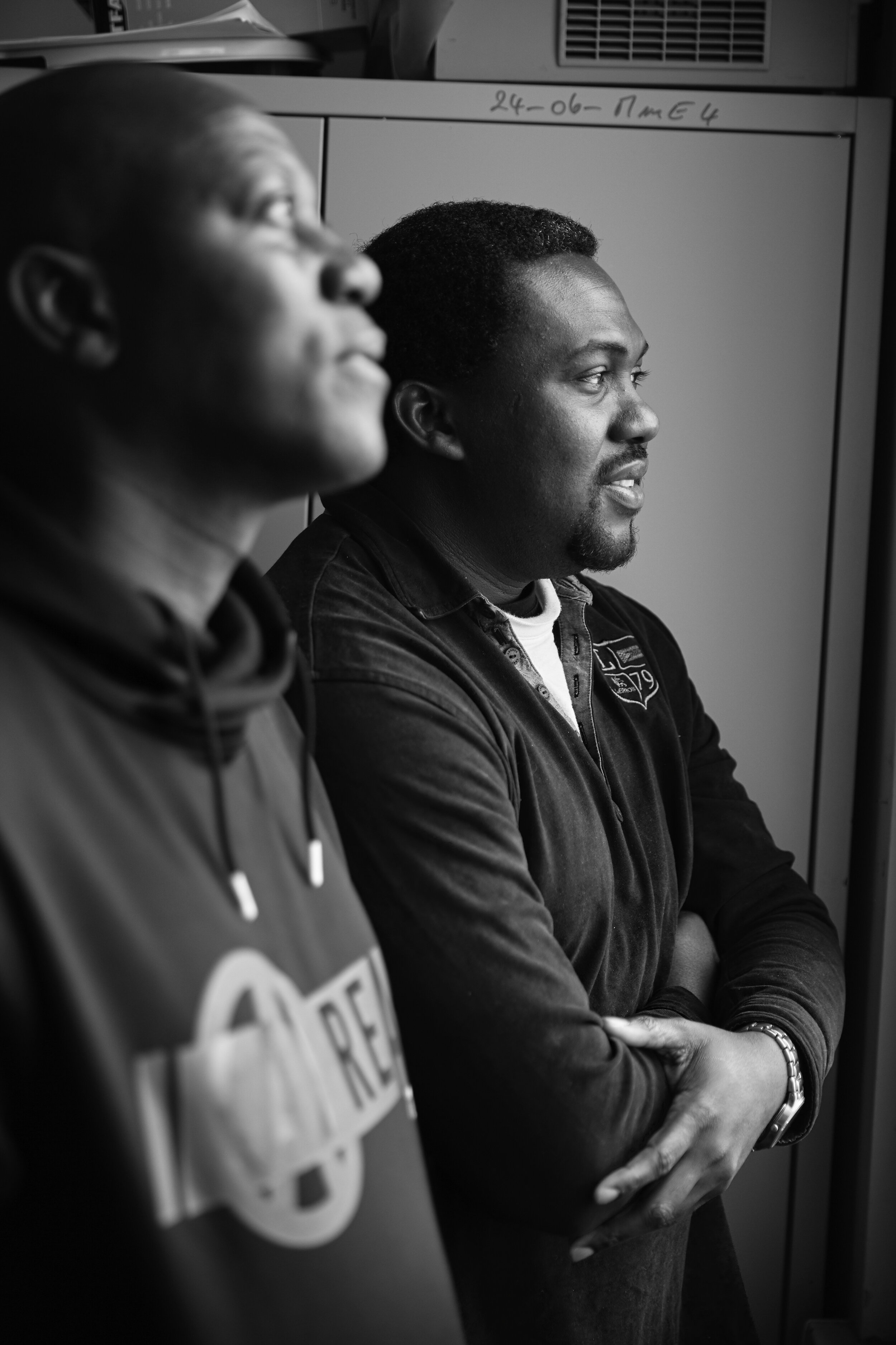 Zwei Männer stehen nebeneinander und blicken zur Seite. Der Mann im Vordergrund trägt einen dunklen Pullover und eine Uhr am Handgelenk. Der Hintergrund zeigt Schränke und Papiere.