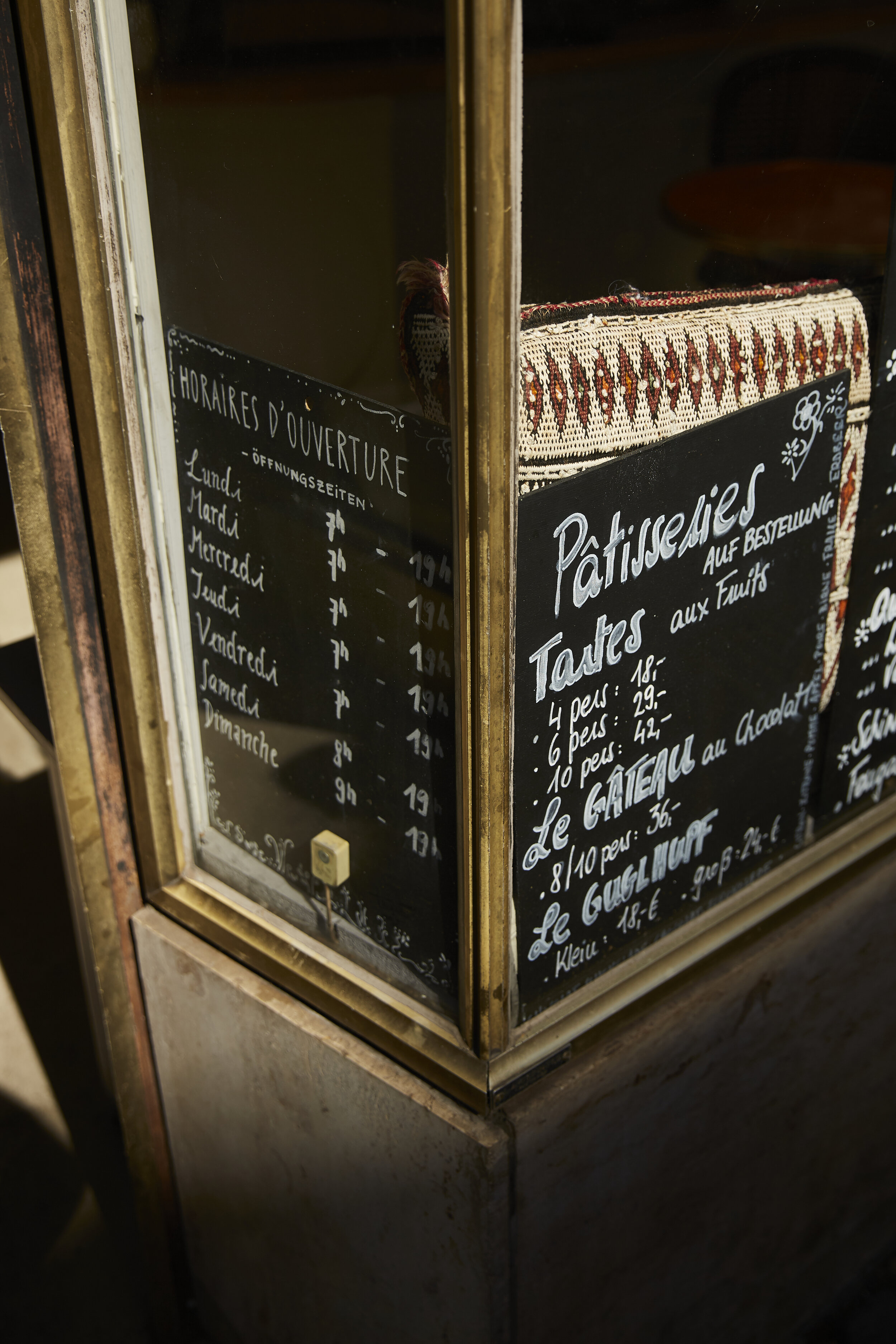 Tafel mit Öffnungszeiten und Patisserie-Angeboten in einem Schaufenster, vermutlich eines Cafés oder einer Bäckerei, mit textlich hervorgehobenen Kuchen und Preisen.