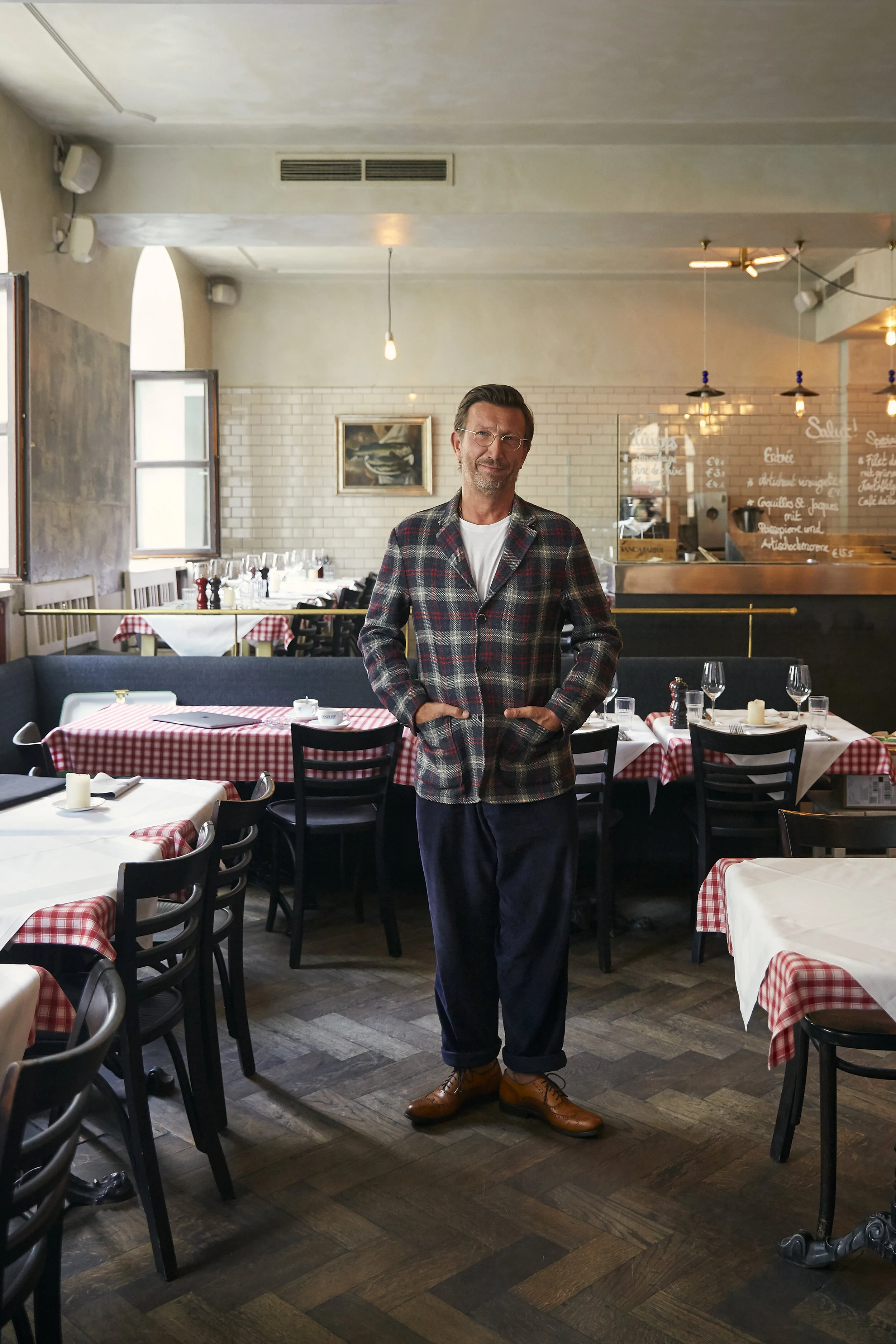 Ein Mann steht in einem Café oder Restaurant mit karierten Tischdecken. Er trägt eine karierte Jacke und Buch. Im Hintergrund sind gedeckte Tische und eine Tafel mit Speisekarte zu sehen.