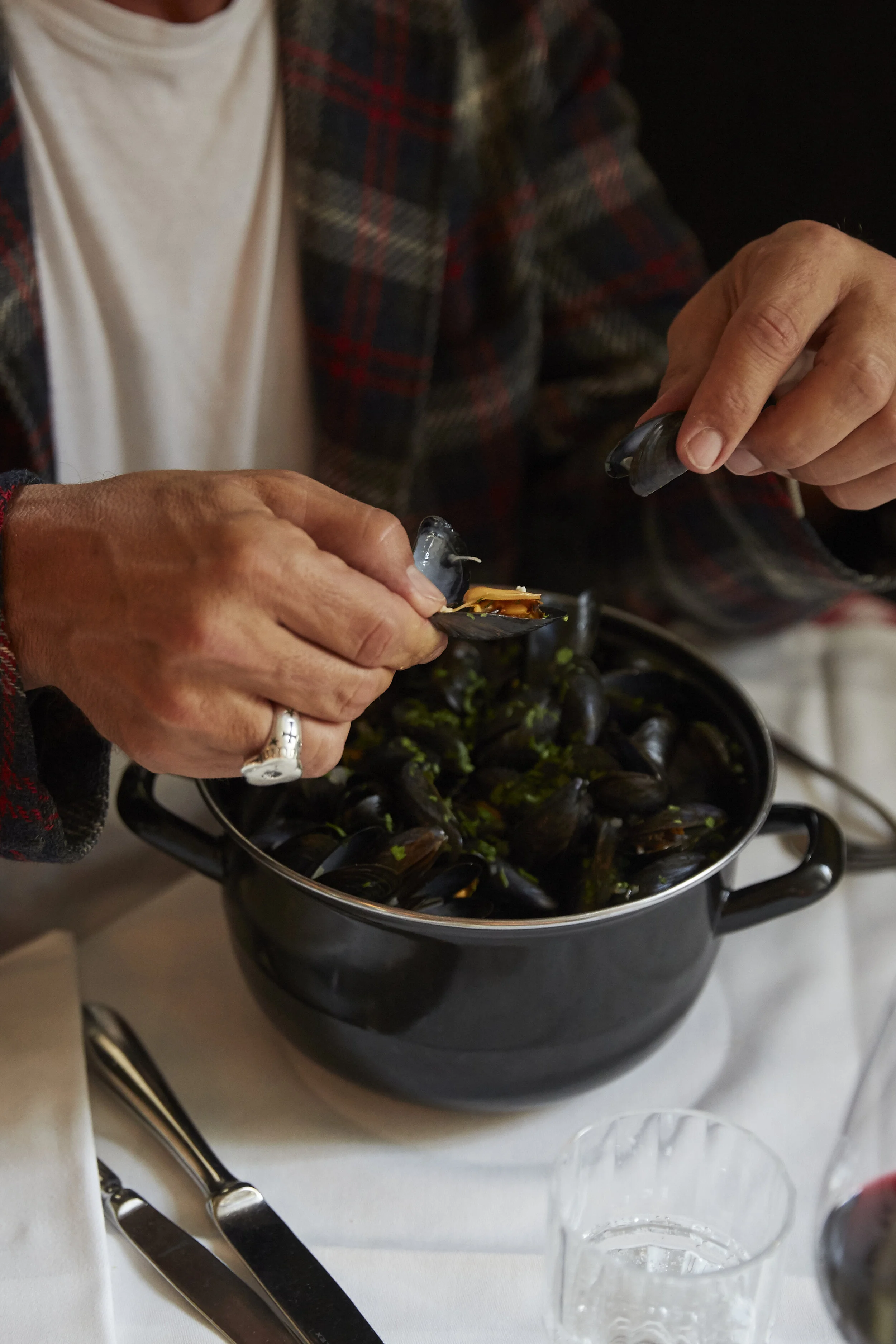 Person isst Miesmuscheln aus einem Topf auf einem Tisch mit Besteck und Glas.