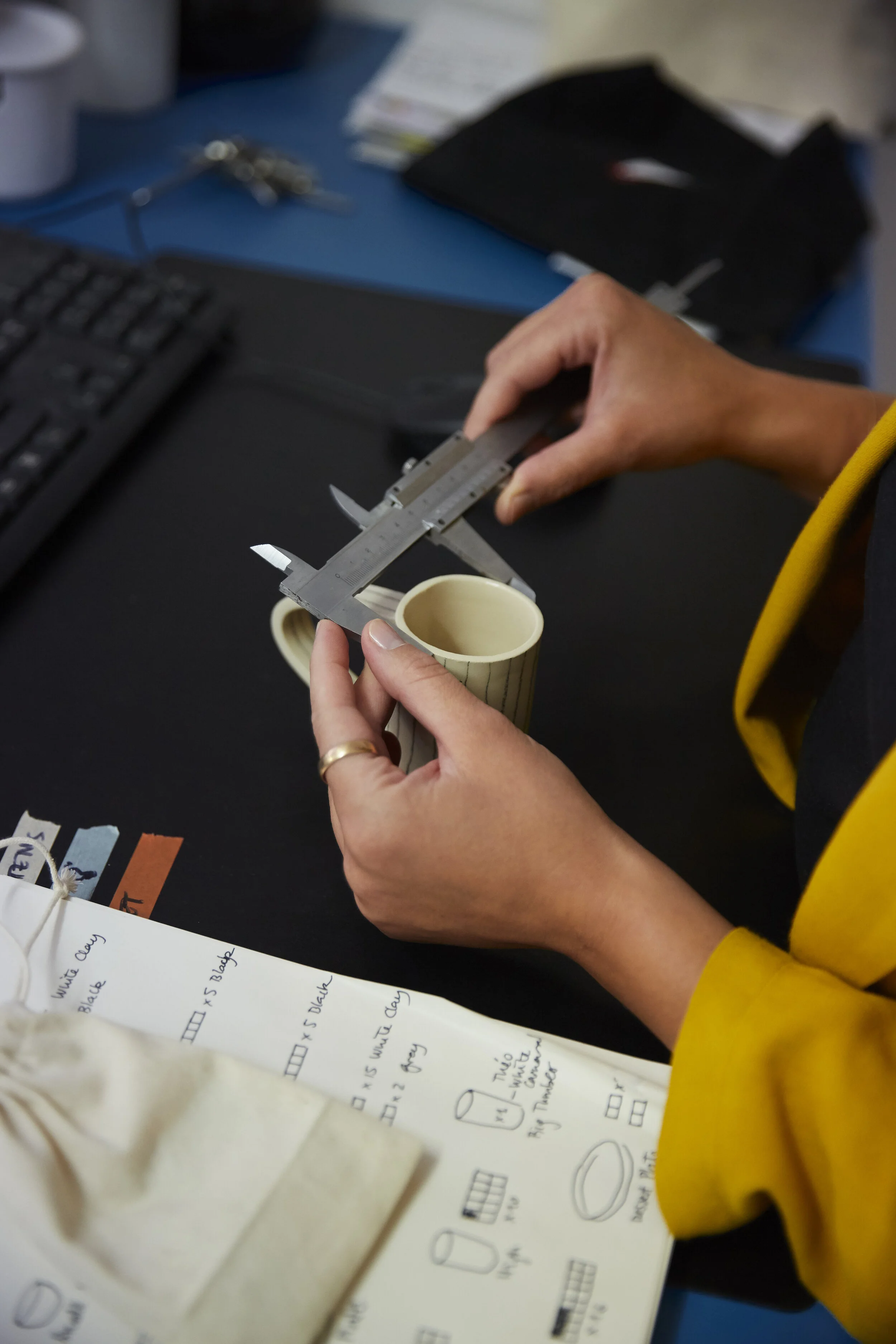 Person misst eine Tasse mit einem Messschieber, Notizbuch im Vordergrund, Tastatur im Hintergrund.