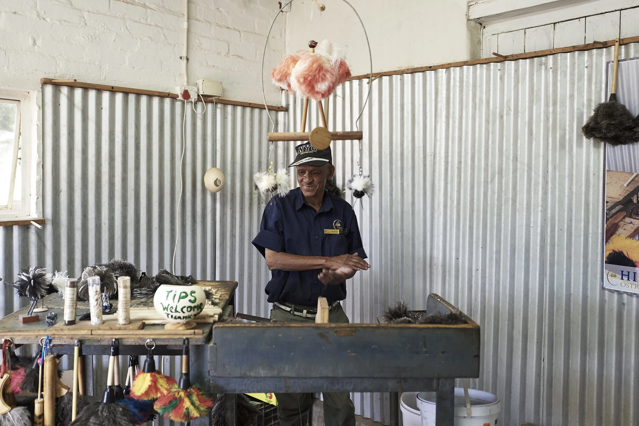 Ein Mann in Uniform steht in einem Raum mit Wellblechwänden. Auf einem Tisch vor ihm liegen verschiedene Staubwedel aus Federn. Ein Schild mit 'Tips Welcome Thank You' ist sichtbar. Im Hintergrund hängen weitere Federprodukte von der Decke.