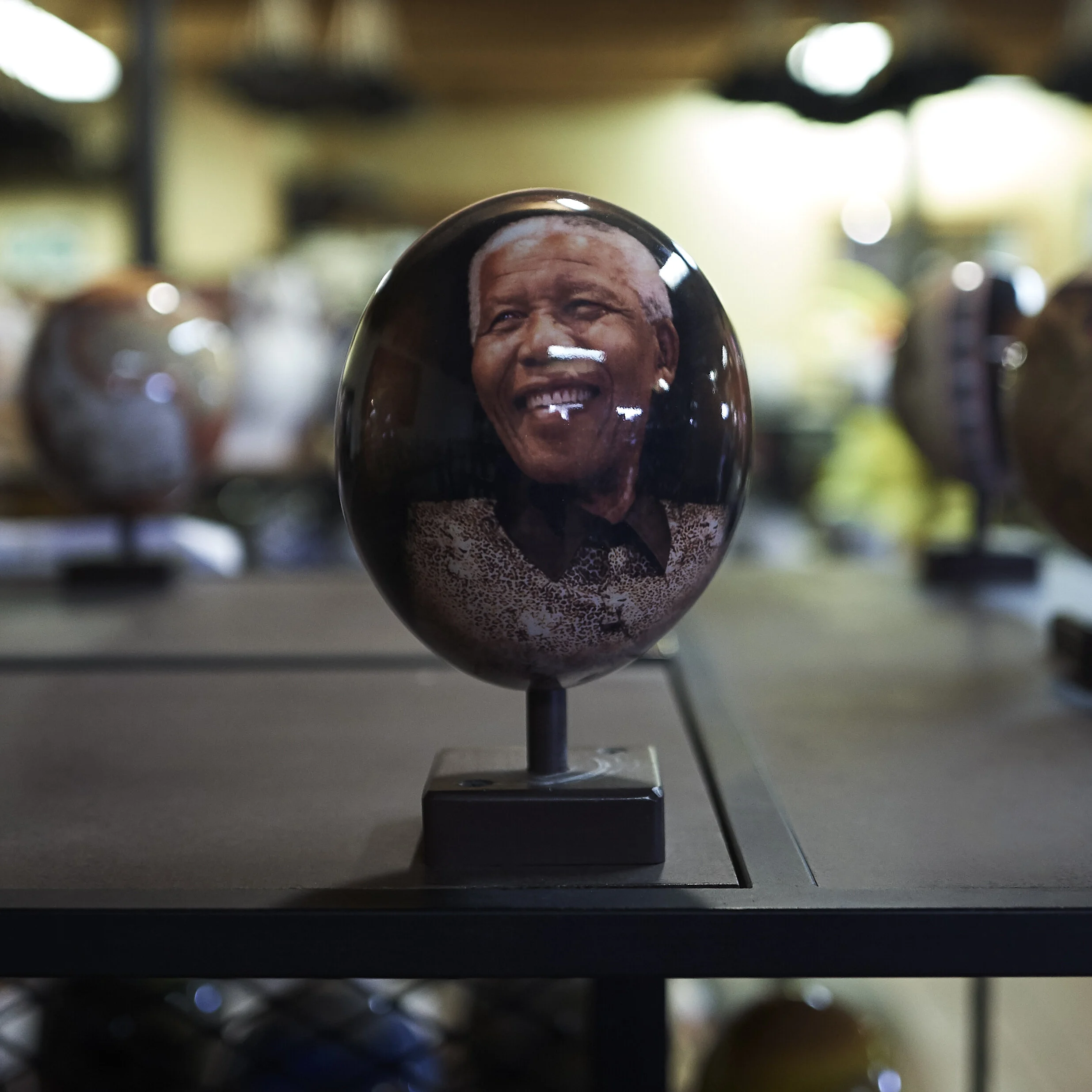 Ein bemaltes Ei mit dem Porträt eines lächelnden Mannes, ausgestellt auf einem Tisch in einem Raum mit unscharfem Hintergrund.