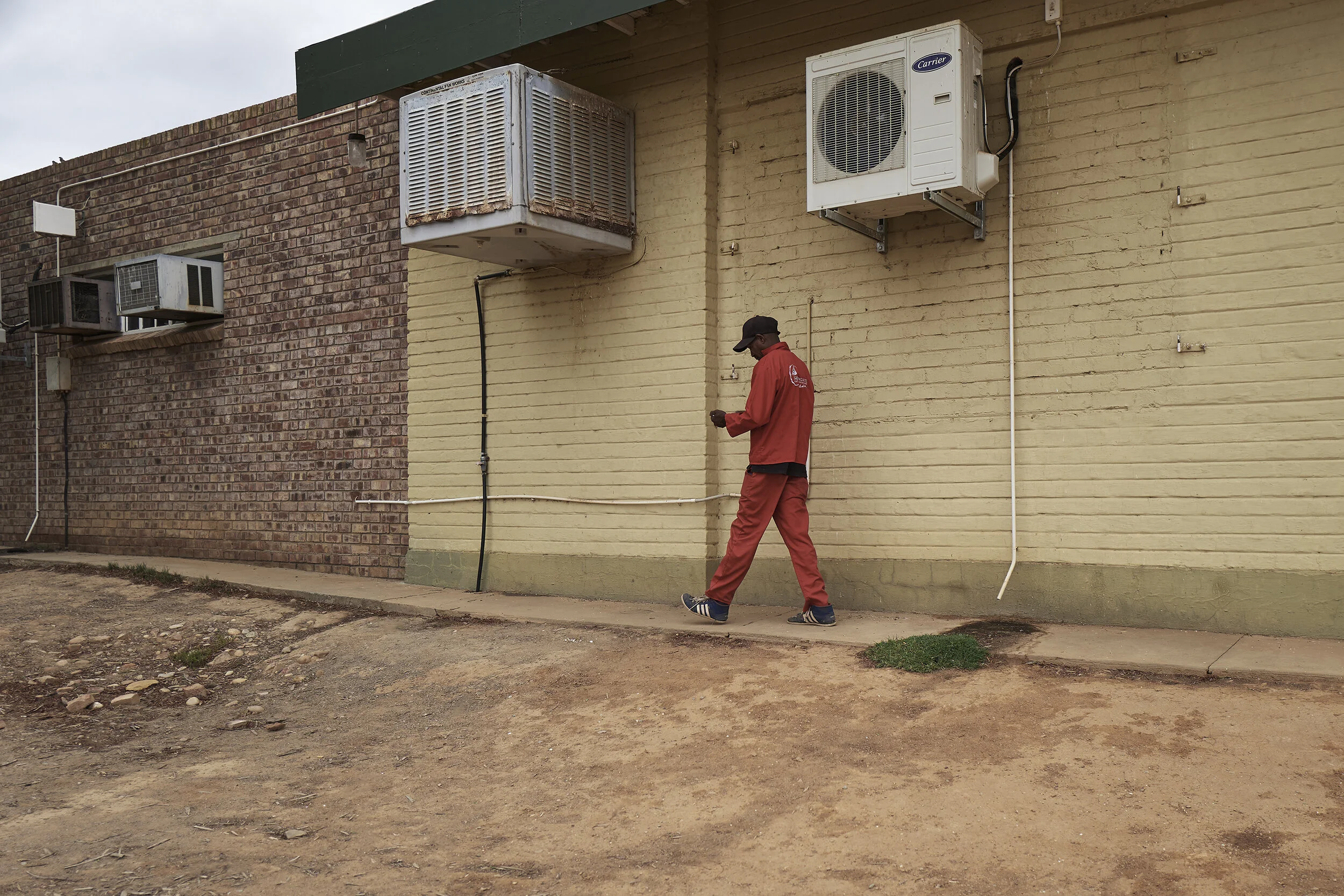 Eine Person in roter Arbeitskleidung läuft an einer Gebäudewand entlang. An der Wand sind mehrere Klimageräte installiert. Der Boden besteht aus sandiger Erde.