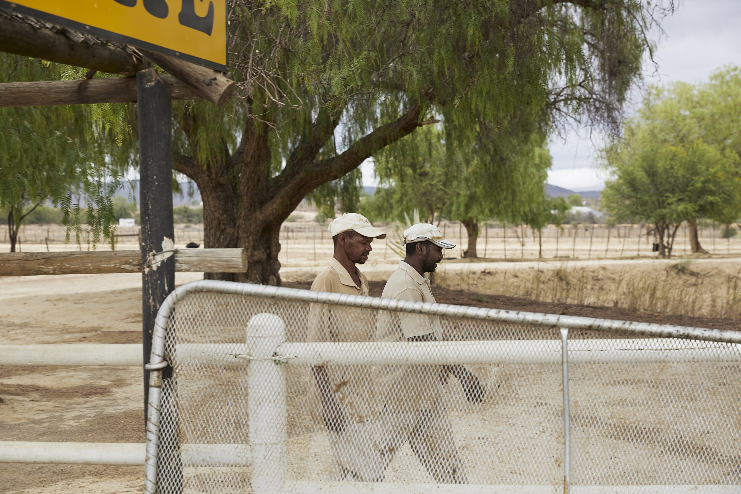 Zwei Männer in beige Kleidung gehen neben einem Zaun, in einer ländlichen Umgebung mit Bäumen und trockenem Boden.
