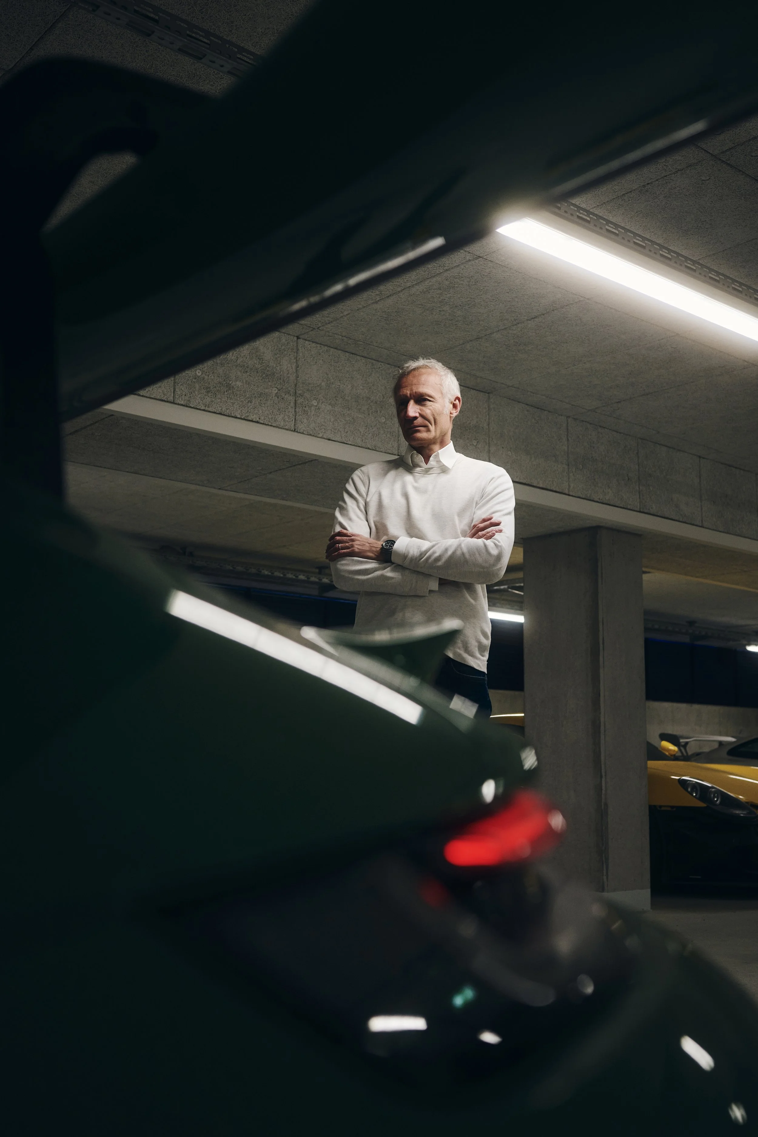 Mann in weißem Pullover steht mit verschränkten Armen in einer Tiefgarage neben einem grünen Auto.