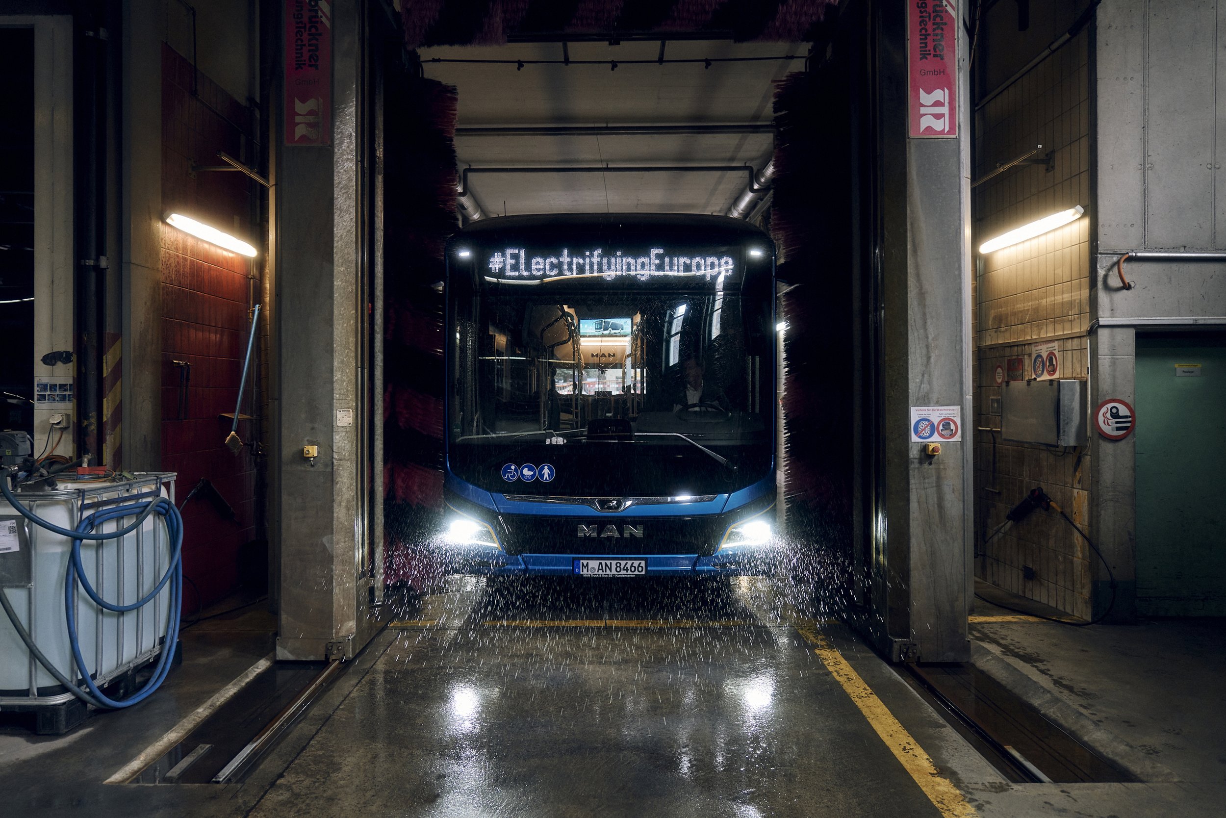MAN-Bus in einer Waschstraße während der Reinigung, mit sichtbar laufendem Wasser und der Aufschrift „#ElectrifyingEurope“ oben.
