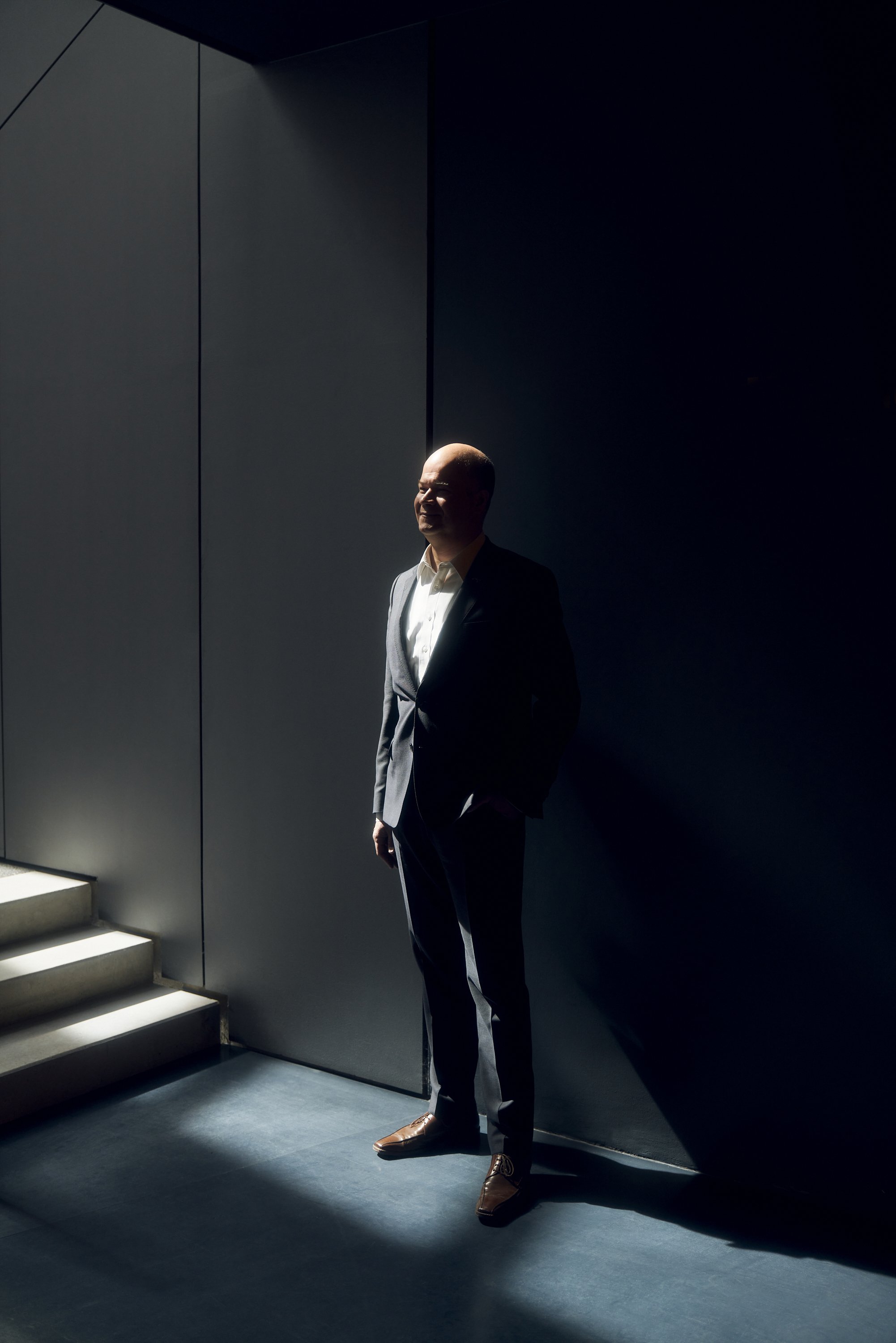 Mann im Anzug steht in einem dunklen Raum, beleuchtet von einem Lichtstrahl, neben einer Treppe.