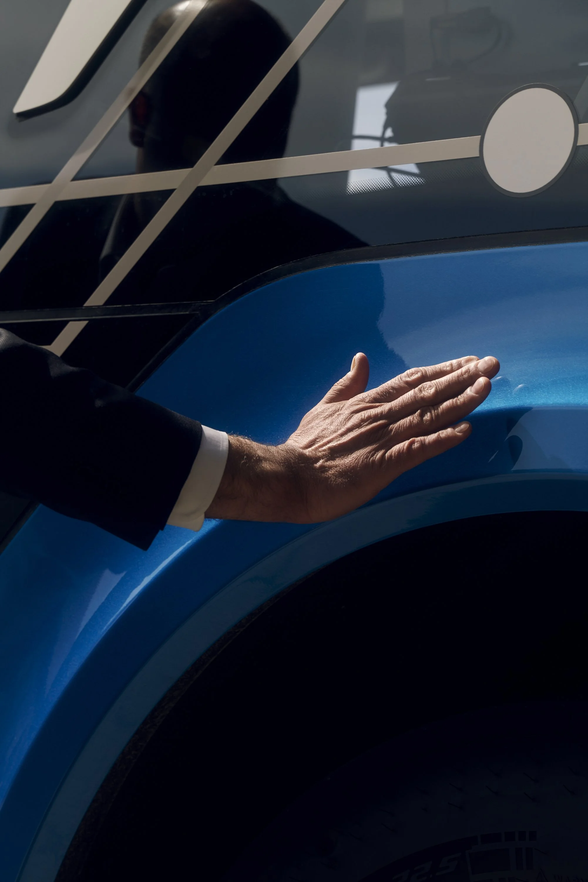Eine Hand berührt die blaue Oberfläche eines Fahrzeugs, reflektierende Linien und das Spiegelbild eines Mannes sind im Hintergrund sichtbar.