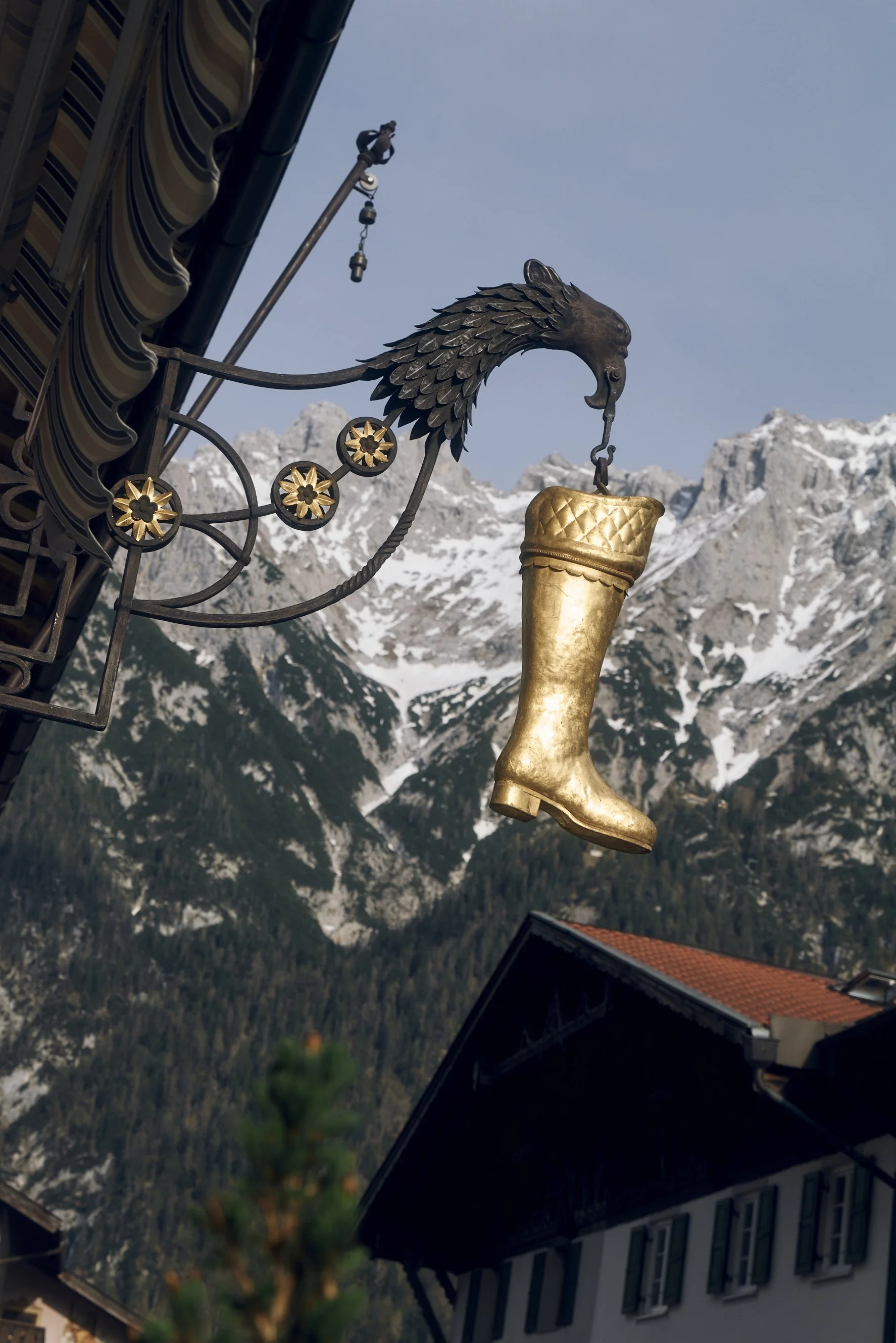Goldener Stiefel an einem kunstvollen Metallschild, dahinter Berglandschaft mit schneebedeckten Gipfeln.