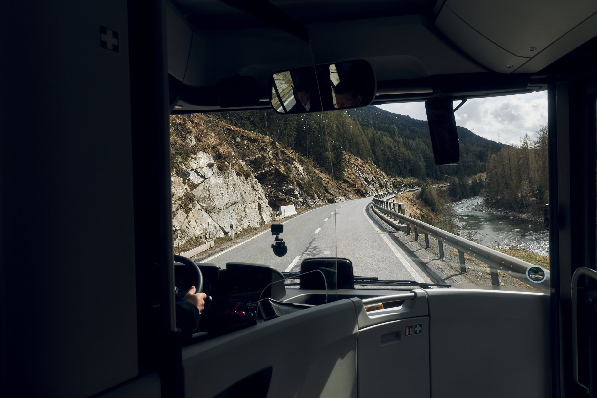 Sicht aus der Fahrerkabine eines Busses auf eine kurvige Bergstraße entlang eines Flusses, umgeben von Felsen und Wäldern.