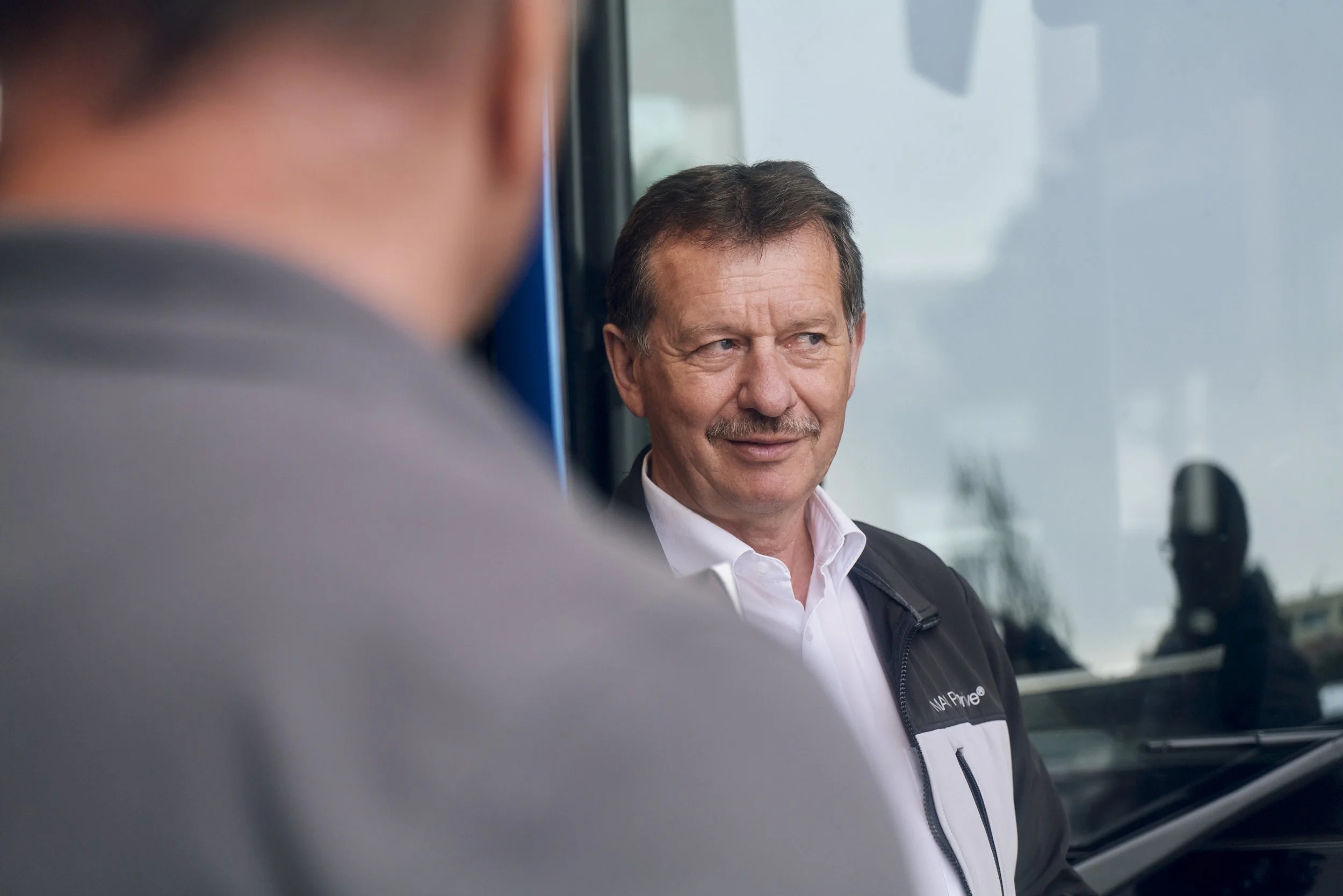 Mann mit Schnurrbart in weißem Hemd und dunkler Jacke, im Gespräch mit einer anderen Person, unscharf im Vordergrund.