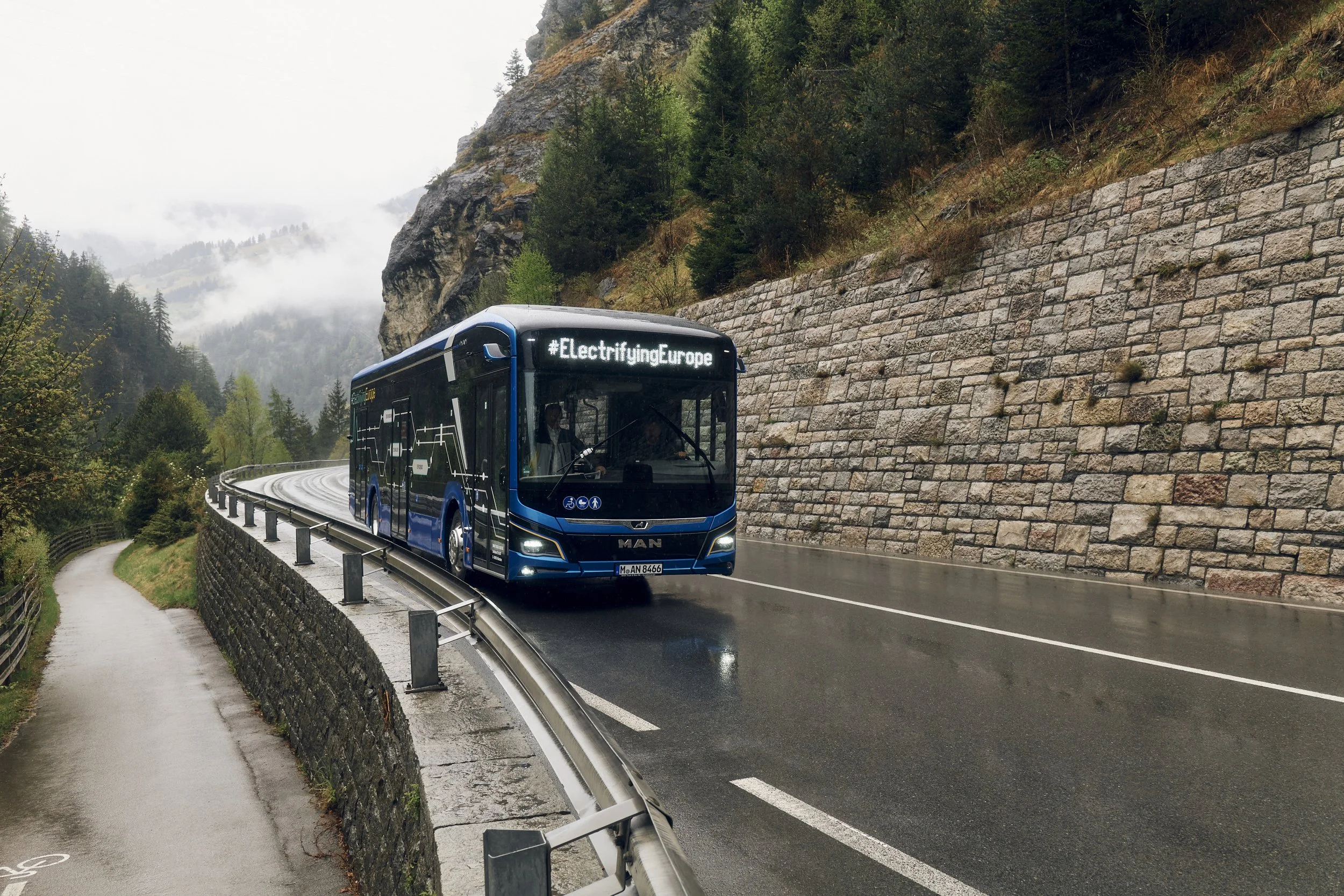 Ein blauer Elektrobus mit der Aufschrift "#ElectrifyingEurope" fährt auf einer regennassen Straße in einer bergigen Landschaft mit Herbstwäldern. Eine Mauer und ein Fahrradweg sind sichtbar.