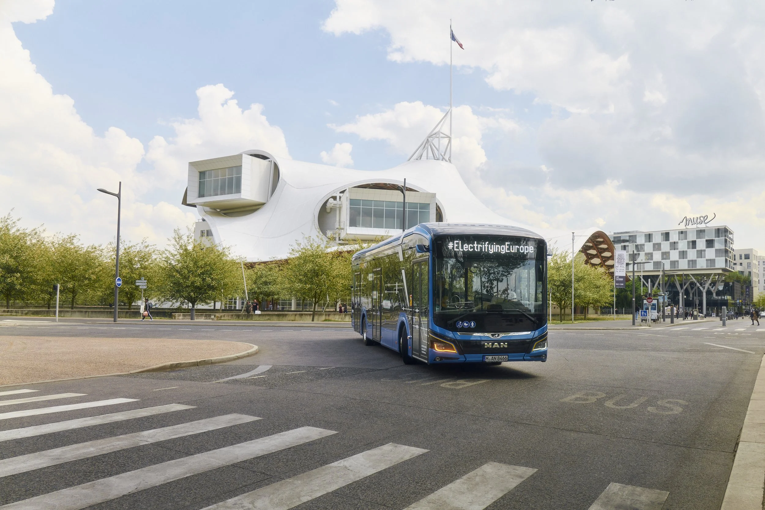 Blauer Bus mit dem Schriftzug #ElectrifyingEurope auf einer Straße vor einem modernen Gebäude mit einer auffälligen Dachstruktur, umgeben von Bäumen. Im Hintergrund ist ein weiteres modernes Gebäude mit dem Namen 'Muse' sichtbar.