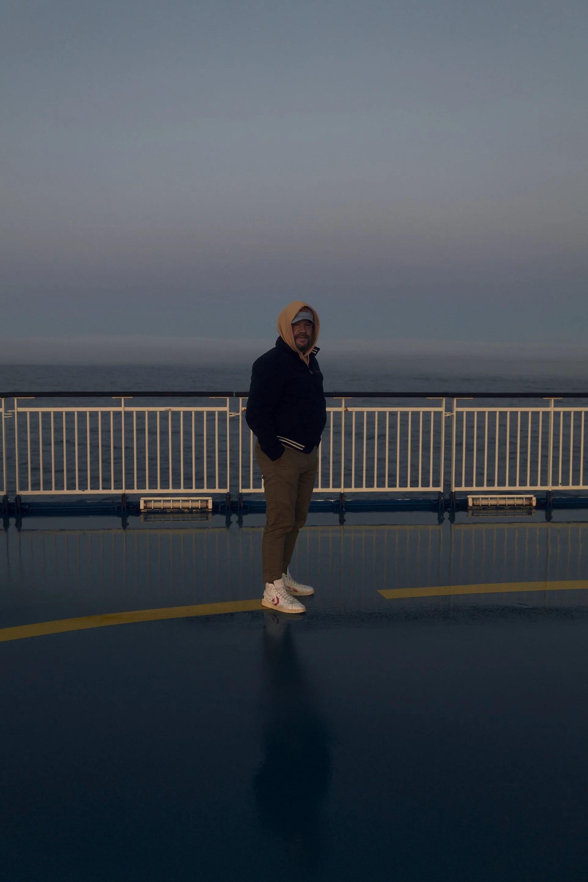 Eine Person in einem Kapuzenpullover steht auf einem Schiffsdeck mit Geländer und Meer im Hintergrund.