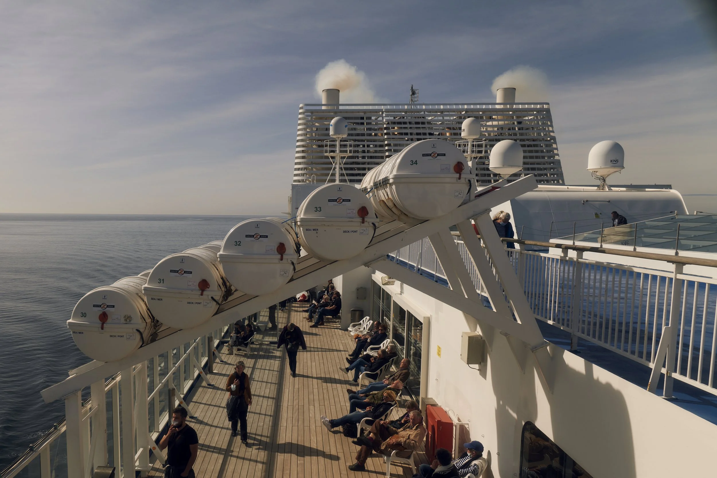 Menschen entspannen sich auf einem Kreuzfahrtschiffdeck mit Rettungsbooten oben und Meereshorizont im Hintergrund.
