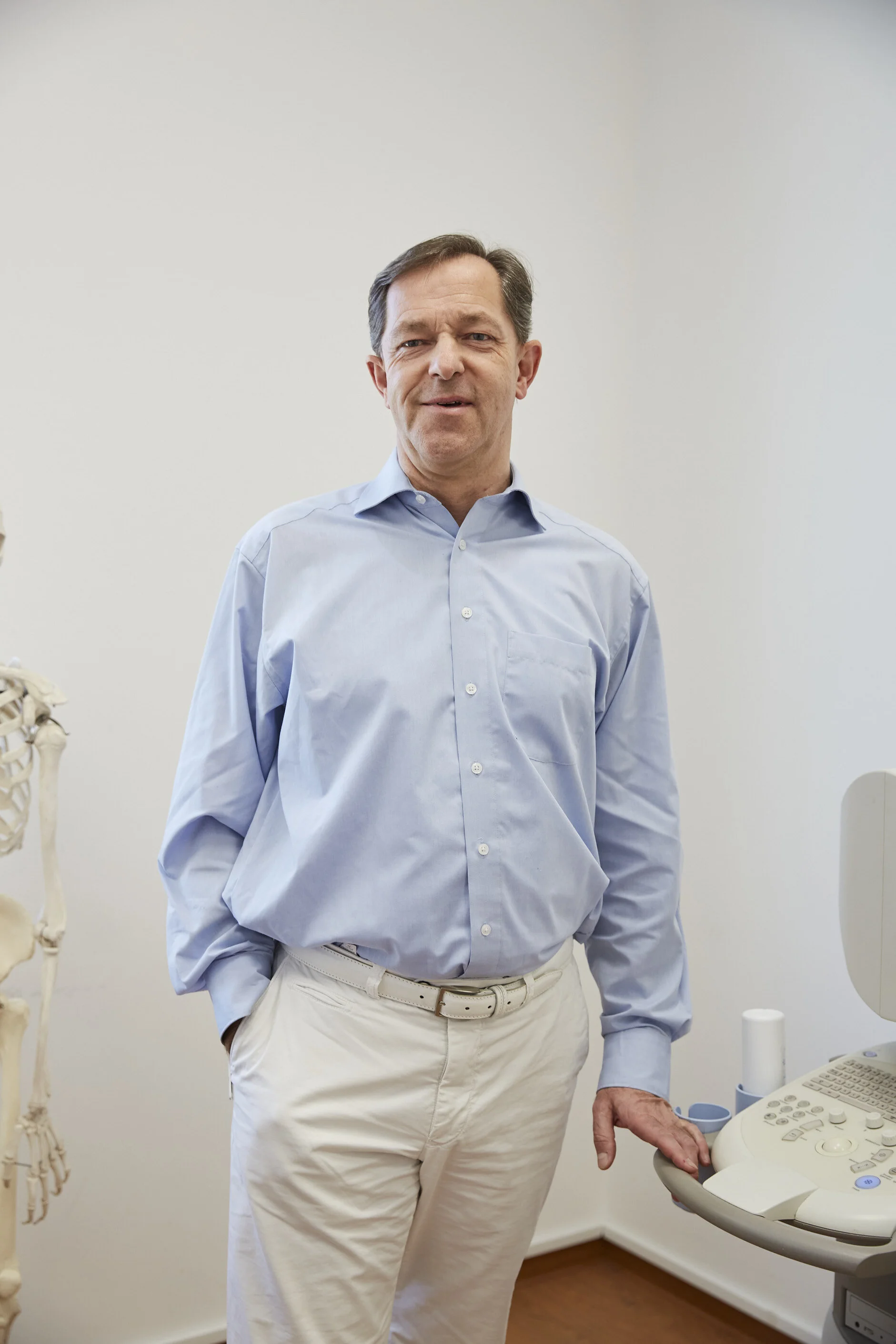 Ein Mann in einem blauen Hemd und weißen Hosen steht in einem Raum neben einem medizinischen Gerät. Links im Bild ist ein Teil eines Skeletts sichtbar.