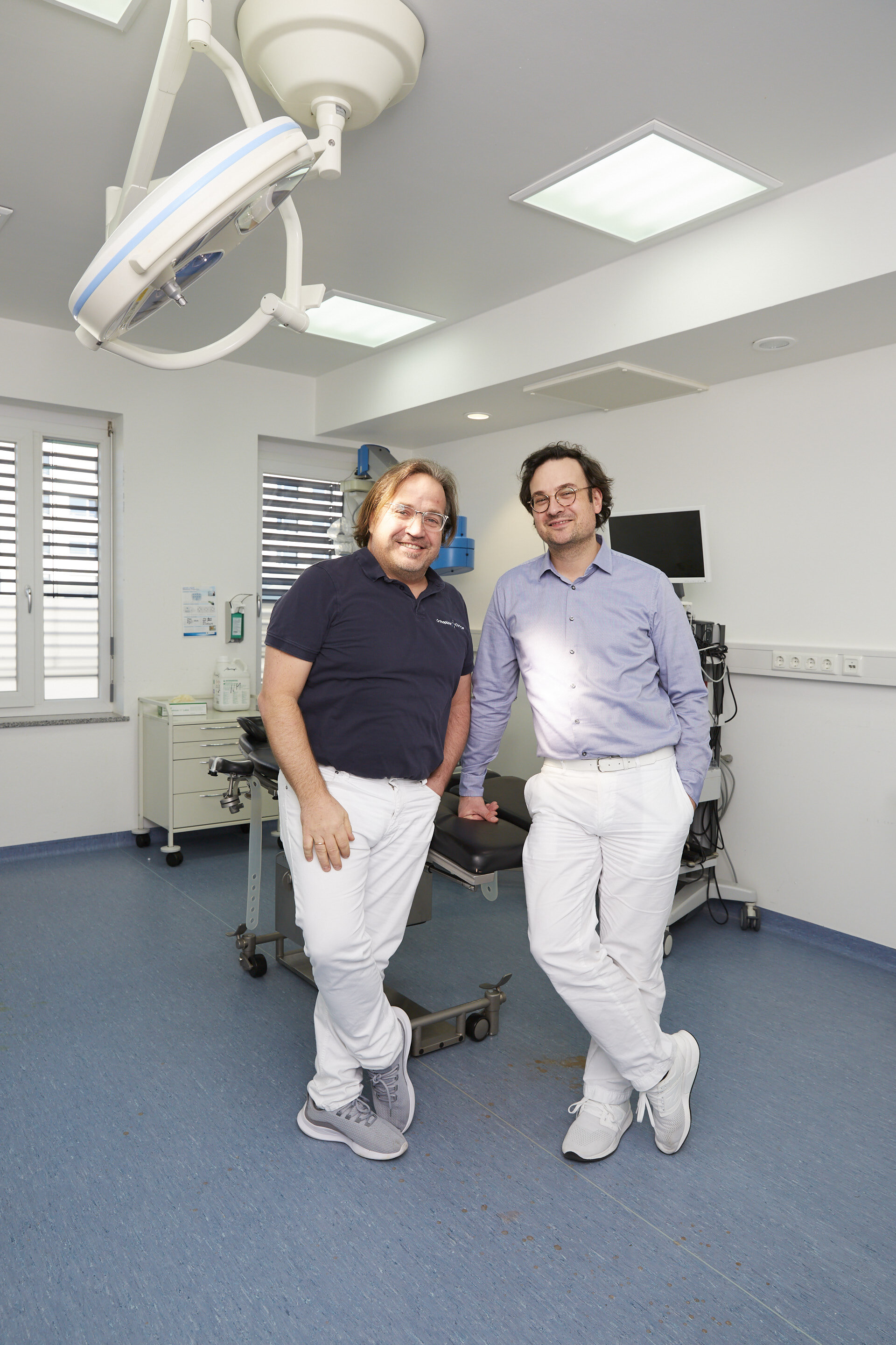 Zwei Männer stehen lächelnd in einem modernen Untersuchungsraum mit medizinischer Ausrüstung und blauem Bodenbelag.