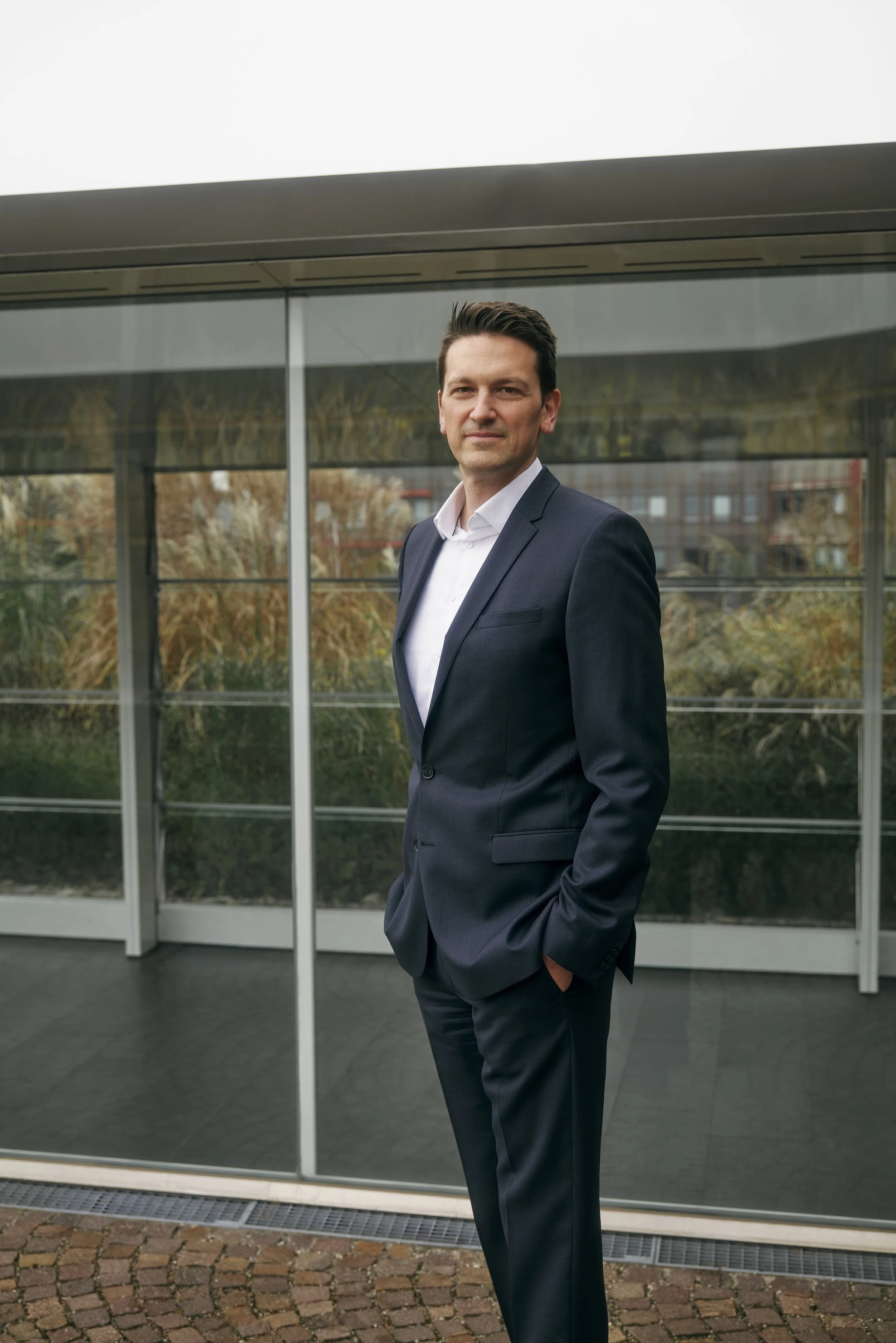 Mann in dunklem Anzug steht vor Glasfassade.