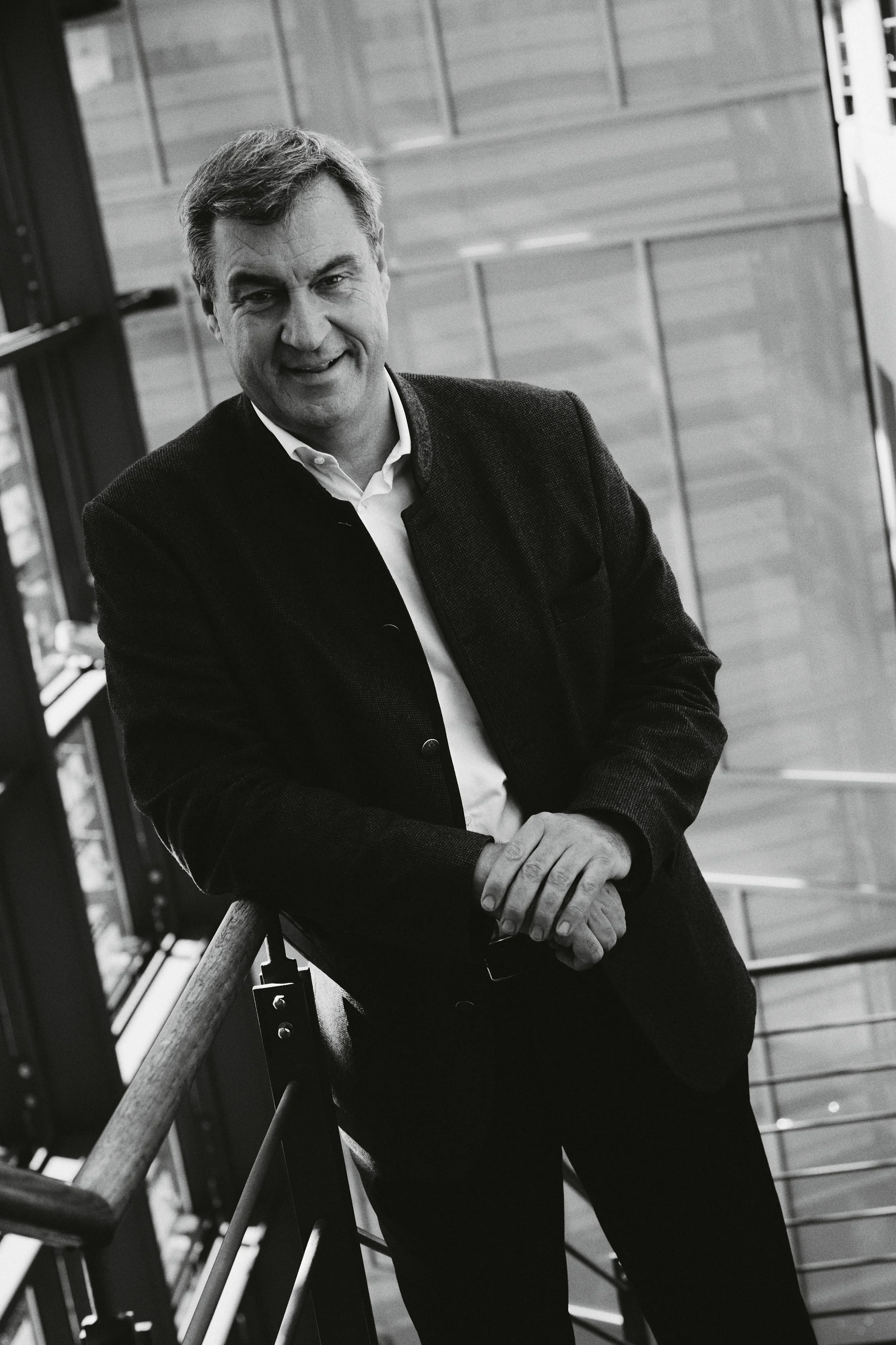 Schwarz-weiß Bild eines lächelnden Markus Söder in einem modernen Gebäude, der sich auf ein Geländer stützt.