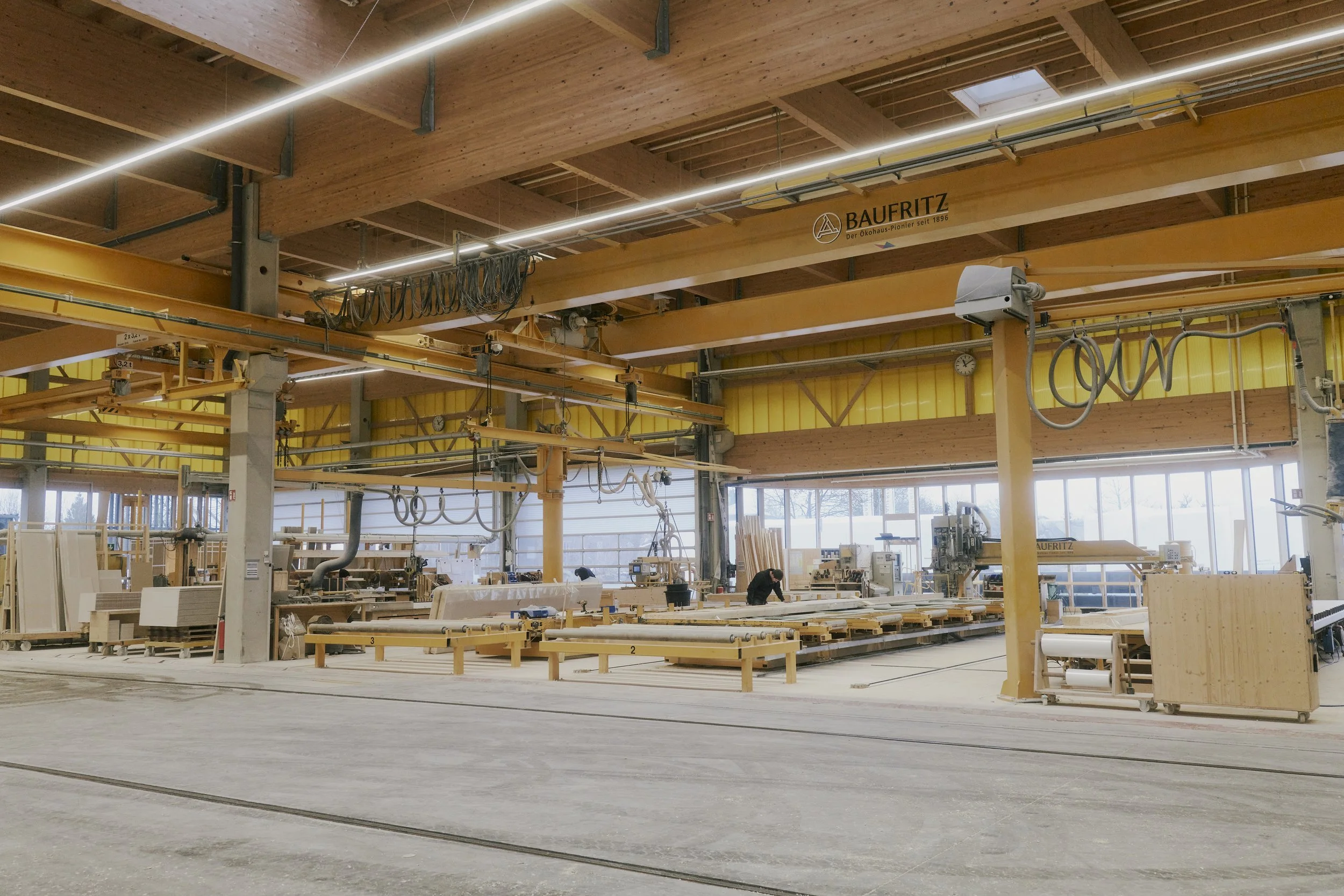 Werkshalle einer Schreinerei mit Holzbearbeitungsmaschinen, Holzplatten und Kränen, modern beleuchtet.