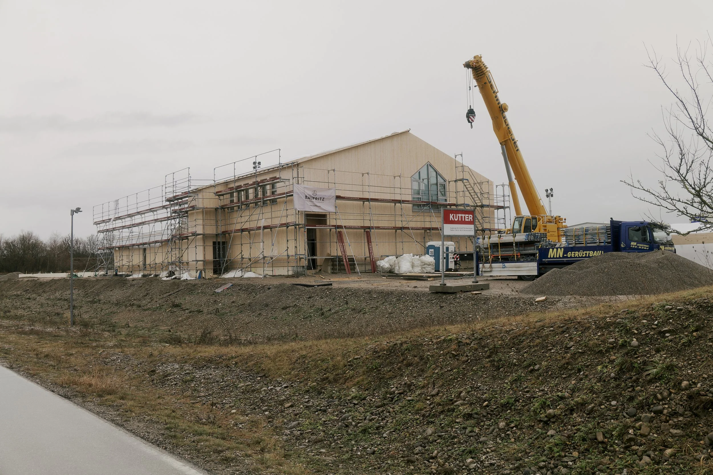 Eine Baustelle mit einem großen Gebäude in der Konstruktion, umgeben von Gerüsten. Ein Kran steht daneben, und ein Lastwagen ist sichtbar. Es gibt einen Hügel aus Erde und Bauschutt, mit einem trüben Himmel im Hintergrund.