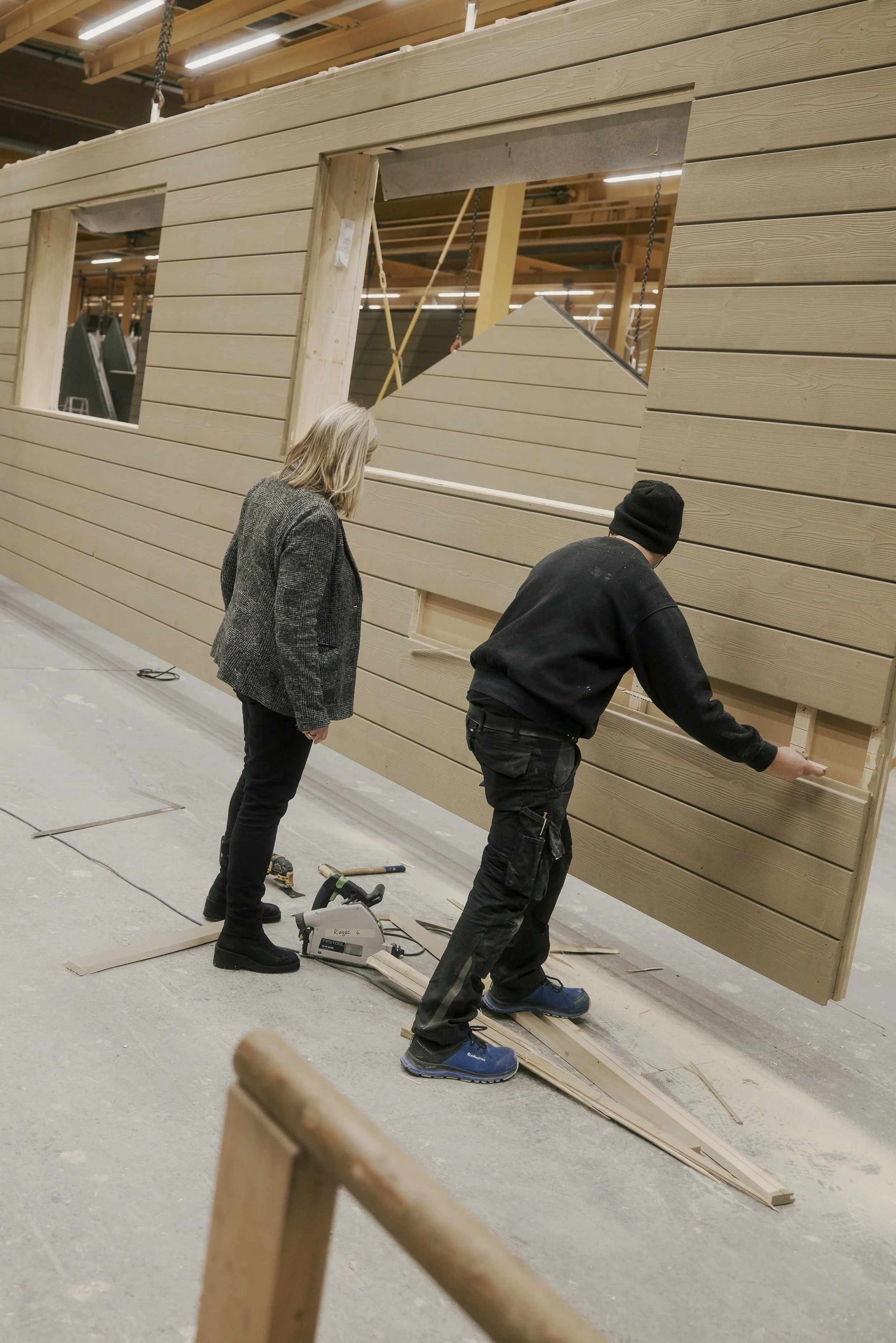 Zwei Personen arbeiten an einer Holzplatte in einer Werkstatt, umgeben von Werkzeugen und Baumaterialien.