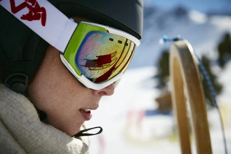 Nahaufnahme einer Person mit Helm und Skibrille, die in einer verschneiten Berglandschaft steht. In der Brille spiegelt sich die Umgebung.