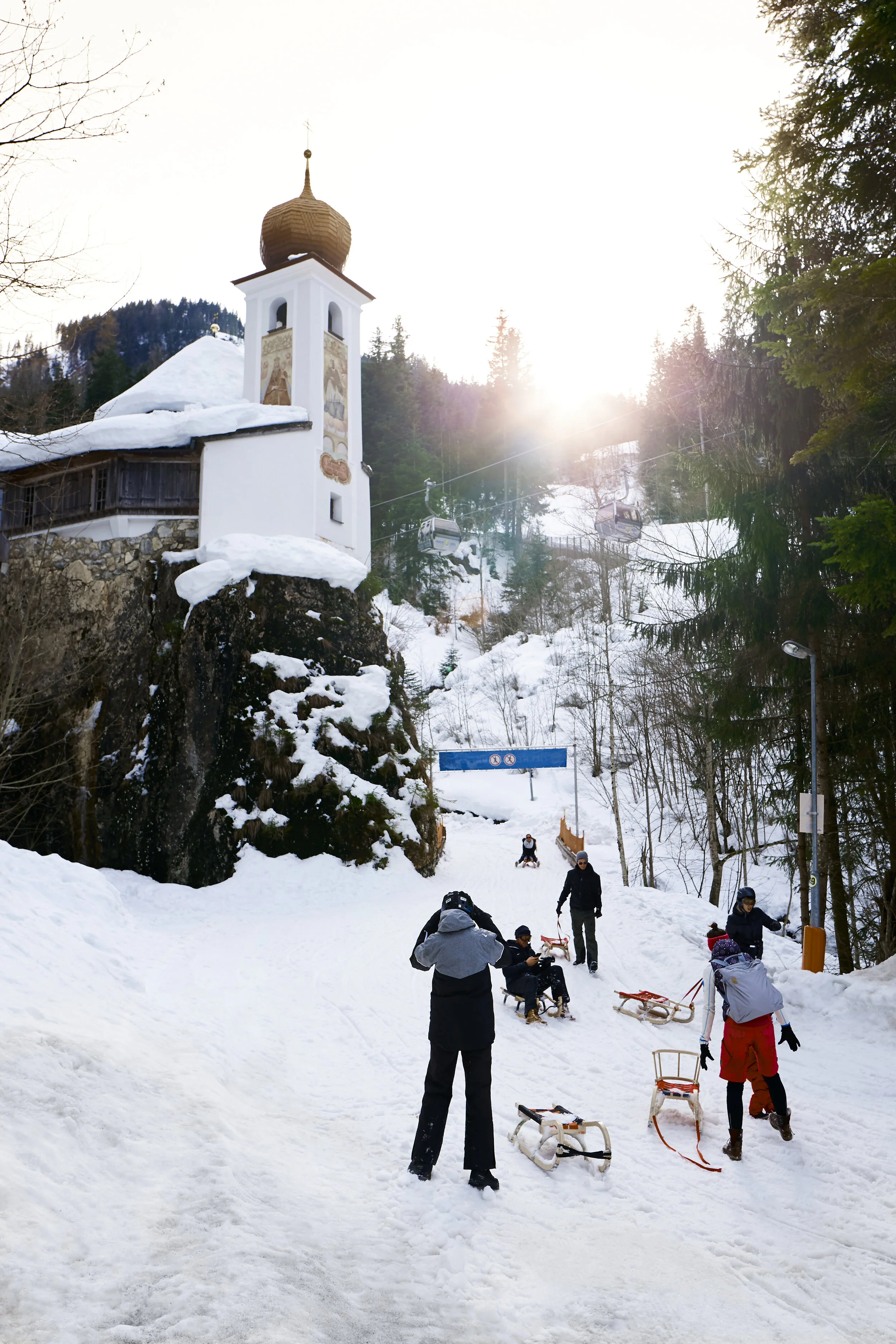 Winterlandschaft mit verschneiter Gebirgskirche, Schlittenfahrern und Wald im Hintergrund. Personen rodeln auf einem verschneiten Pfad.