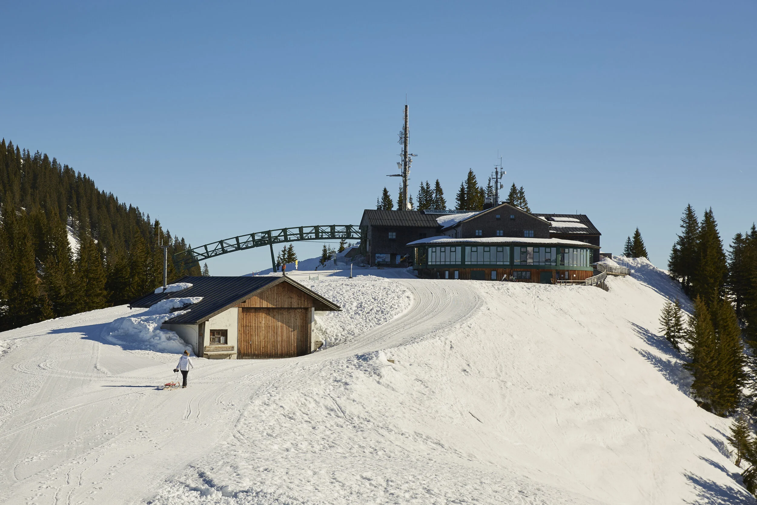 Verschneite Berglandschaft mit einem Gebäude auf einem Hügel, umgeben von Nadelbäumen, unter blauem Himmel. Ein einzelner Mensch zieht einen Schlitten über den Schnee.