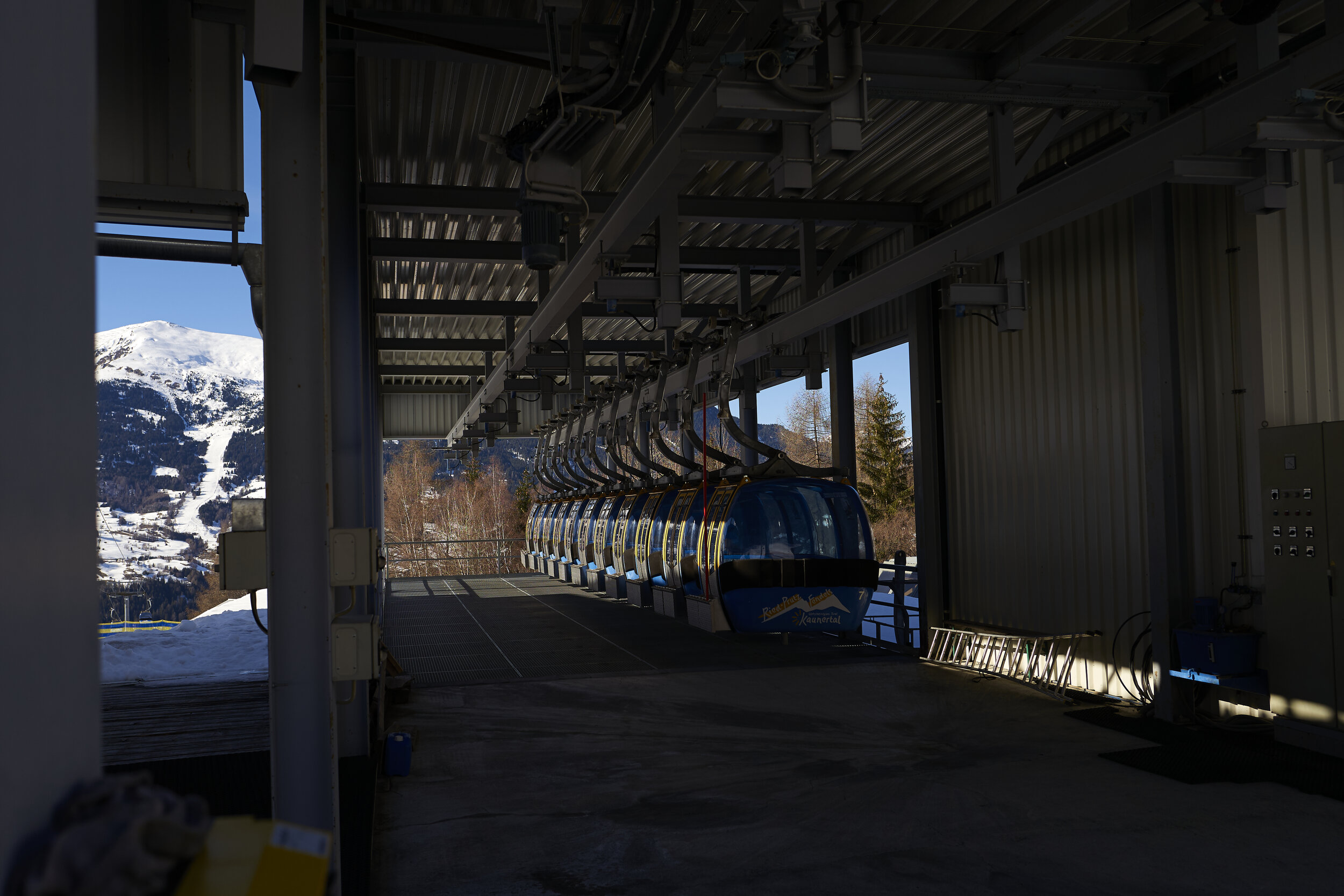 Seilbahnstation mit Gondeln, Bergpanorama, Schnee bedeckte Berge im Hintergrund, Stahlkonstruktion, Winter, bewaldete Hanglandschaft.
