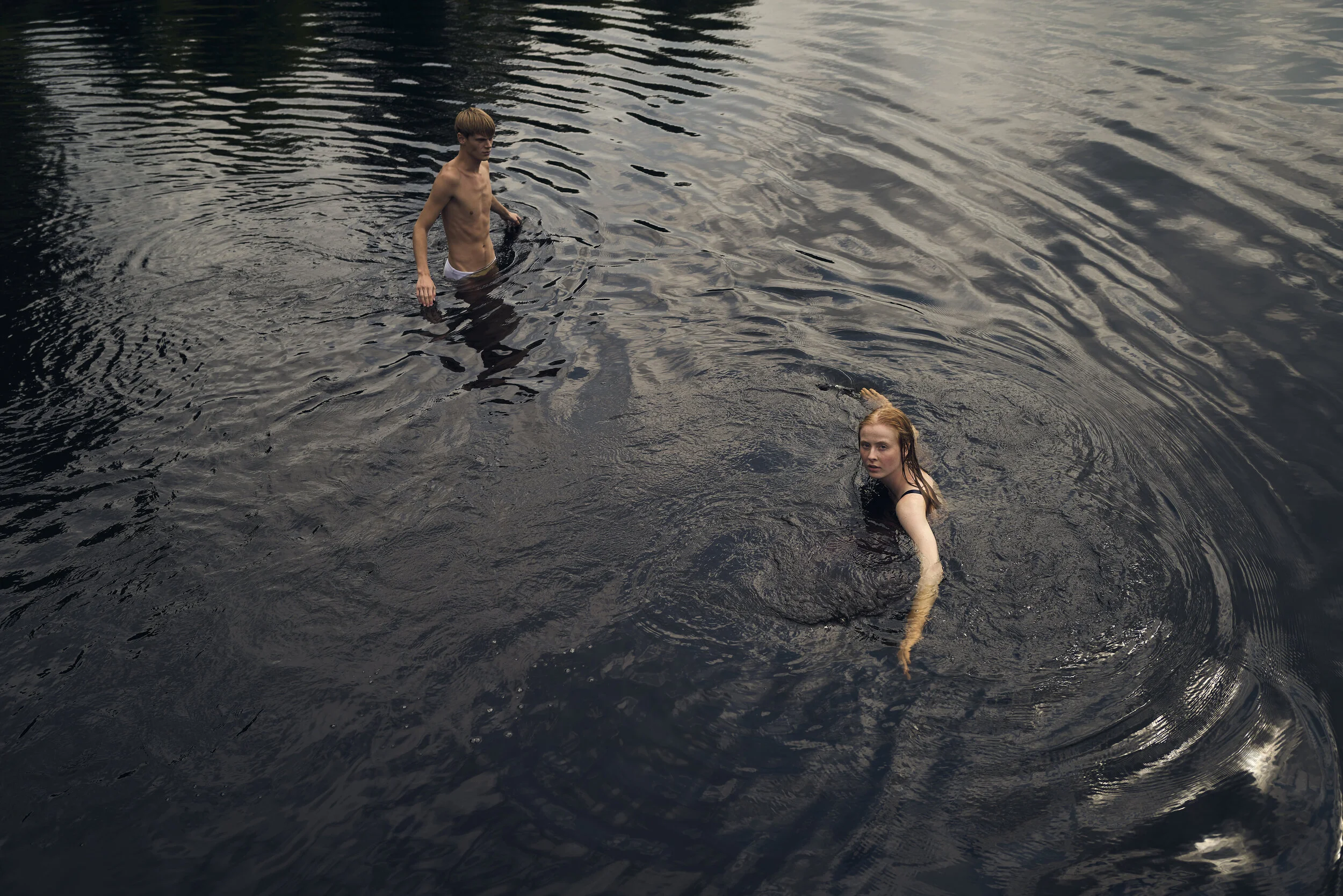 Zwei Personen schwimmen in einem dunklen Gewässer. Der Mann steht im Wasser, während die Frau schwimmt und zur Kamera schaut.