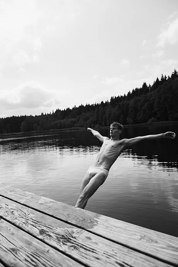 Schwarz-weiß-Foto eines Mannes, der in Unterwäsche von einem Steg in einen See springt. Im Hintergrund ist ein Wald zu sehen.